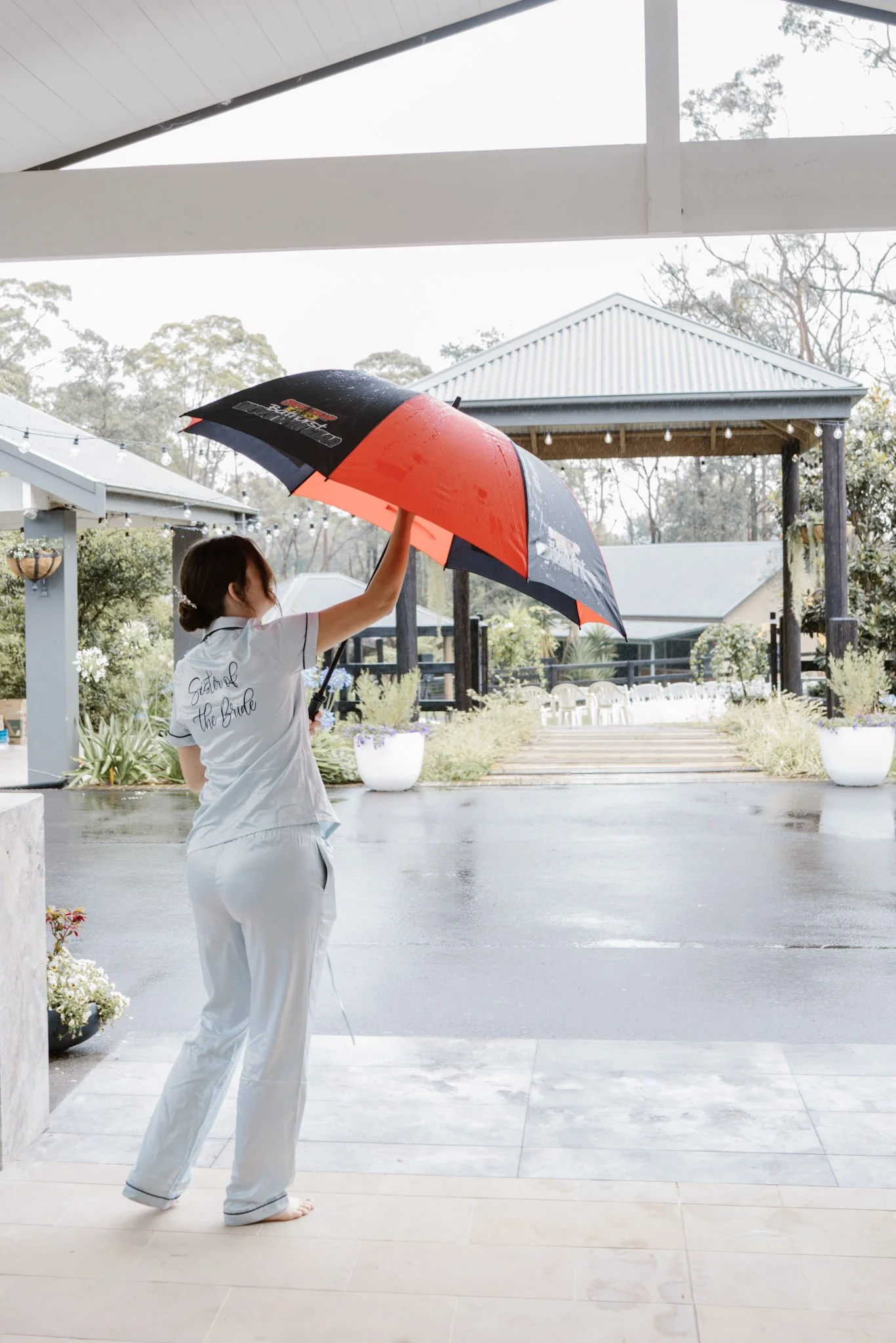 A woman wearing white pajamas with 'Easter the Bride' written on the back, holding a red and black umbrella, standing barefoot on a porch during a rainy day, with a view of a garden and a structure with a peaked roof in the background.