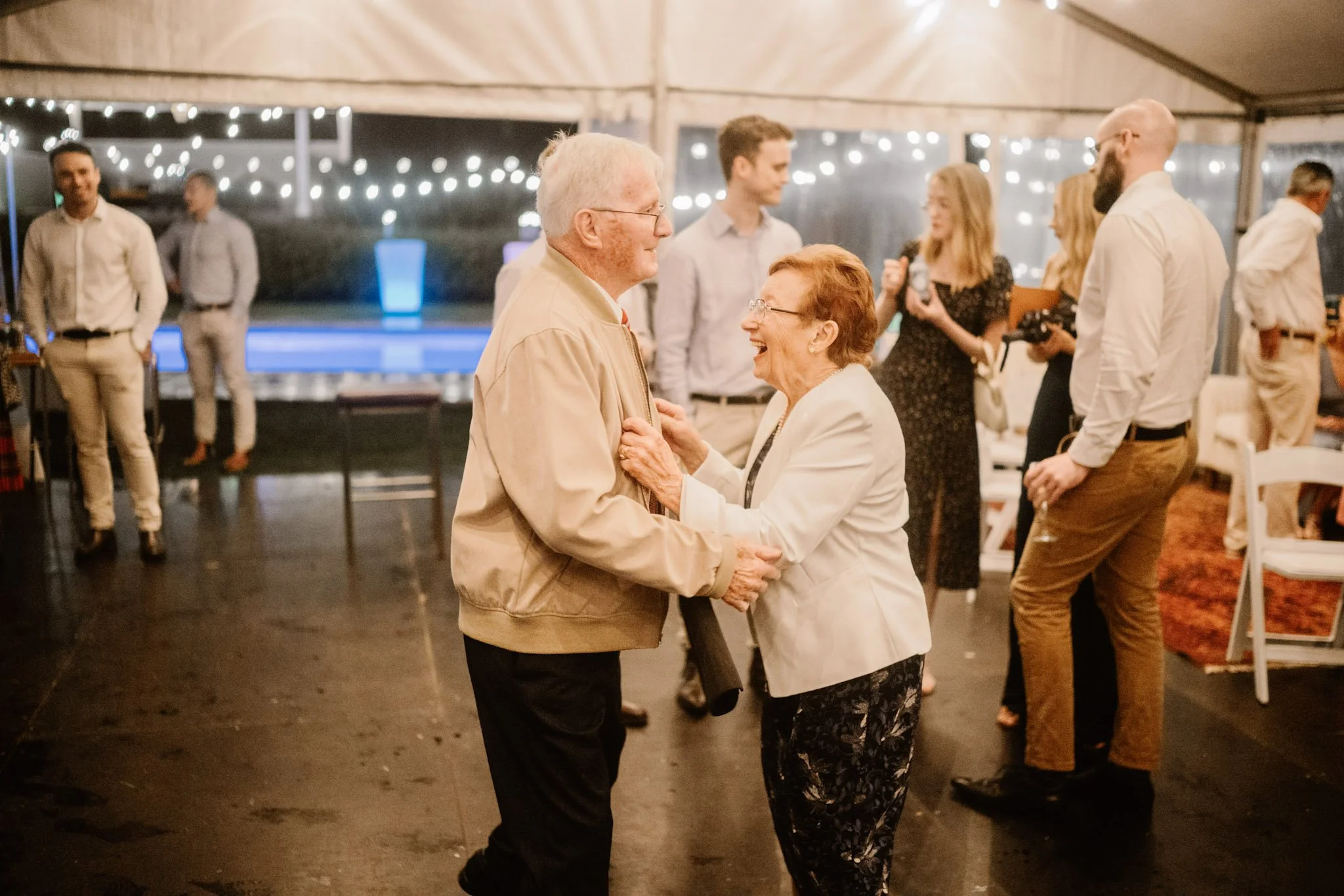 An elderly couple dancing and smiling at a celebration inside a decorated tent, with string lights and several people chatting and enjoying the event.