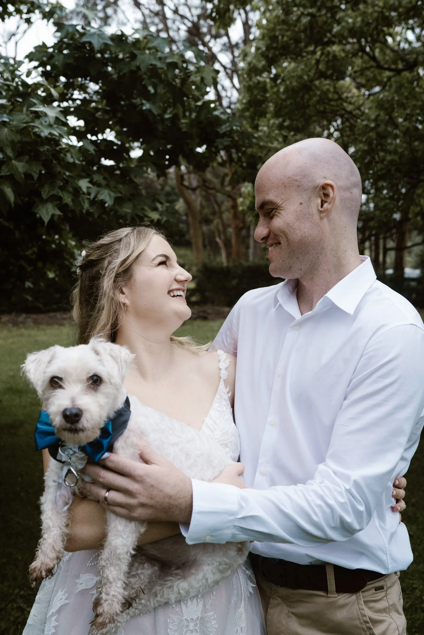 A couple, dressed in wedding attire, smiling and looking at each other while holding a small white dog with a blue bow tie, outdoors with green trees in the background.