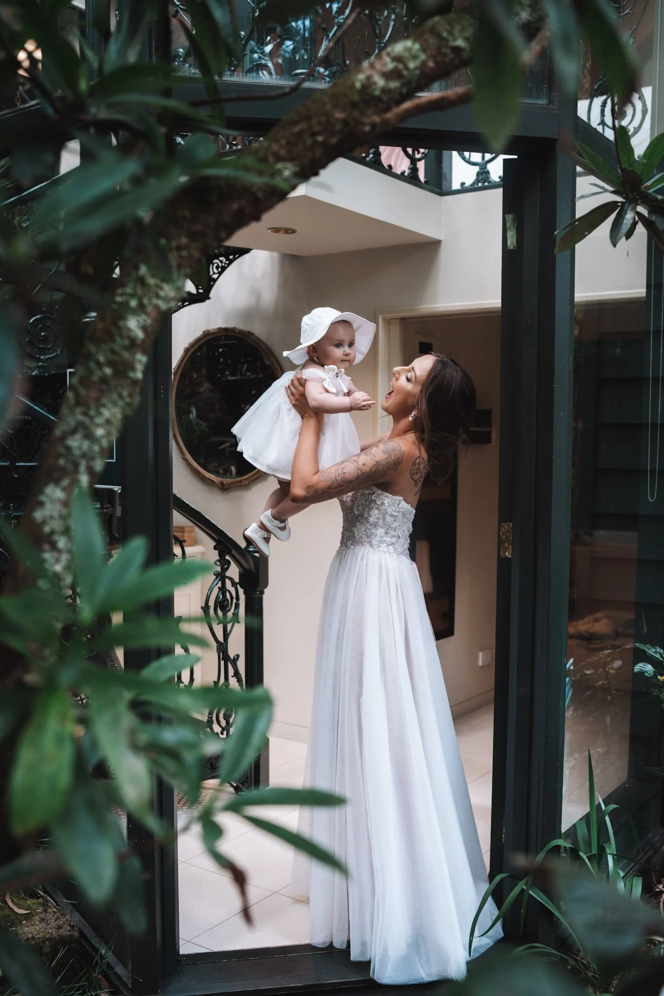 A woman in a white dress holding a young girl in a white dress and hat inside a house, viewed through a glass door with greenery in the foreground.