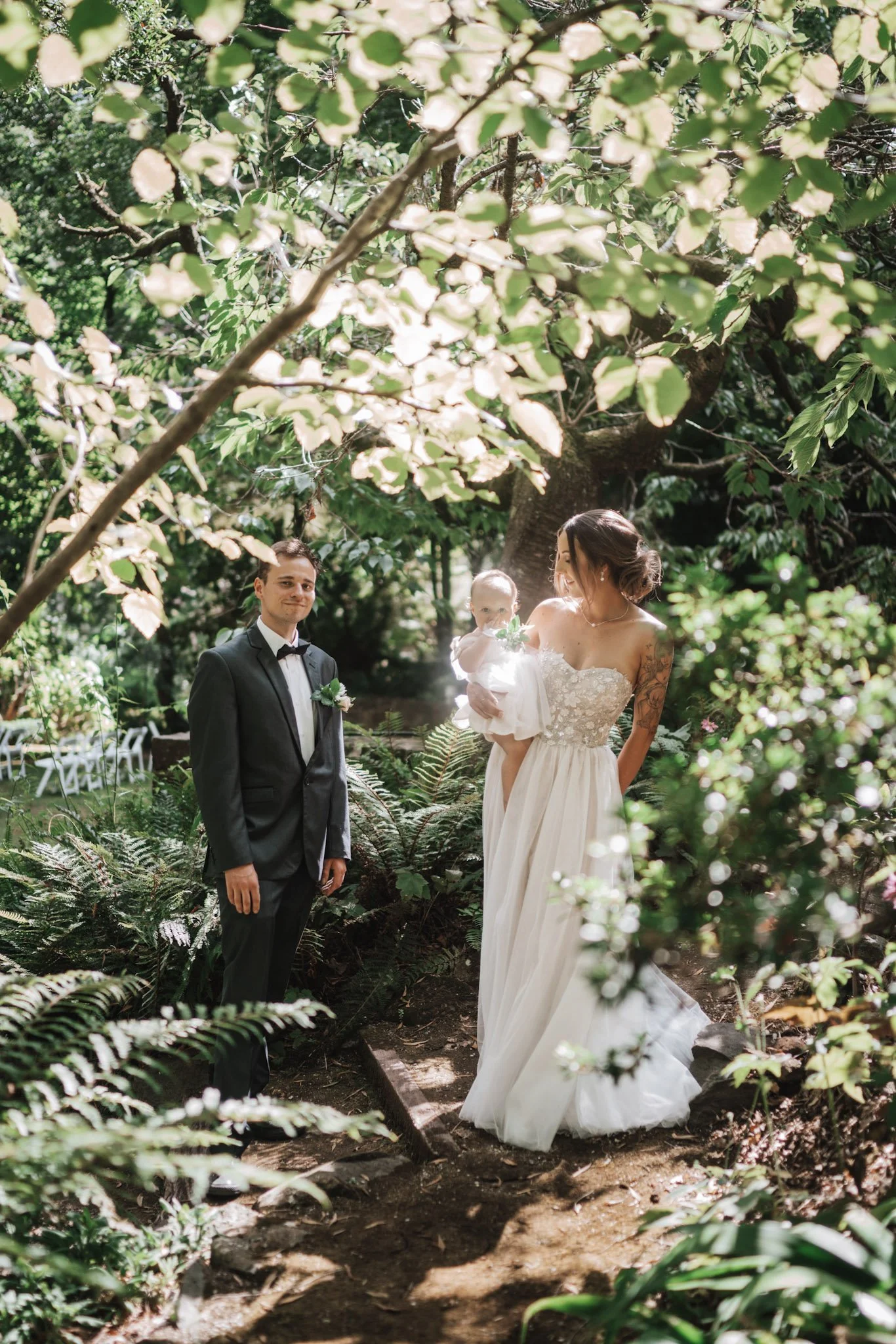 A wedding scene in a lush garden with a bride in a white strapless gown holding a baby in a white dress, a groom in a black tuxedo with a bow tie, surrounded by greenery and trees.