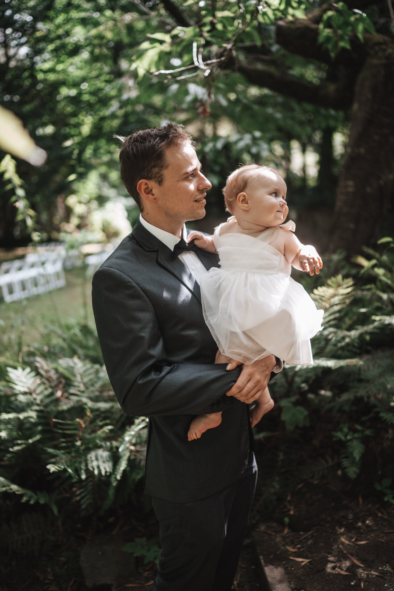 A man in a tuxedo holds a young girl in a white dress outdoors among greenery.