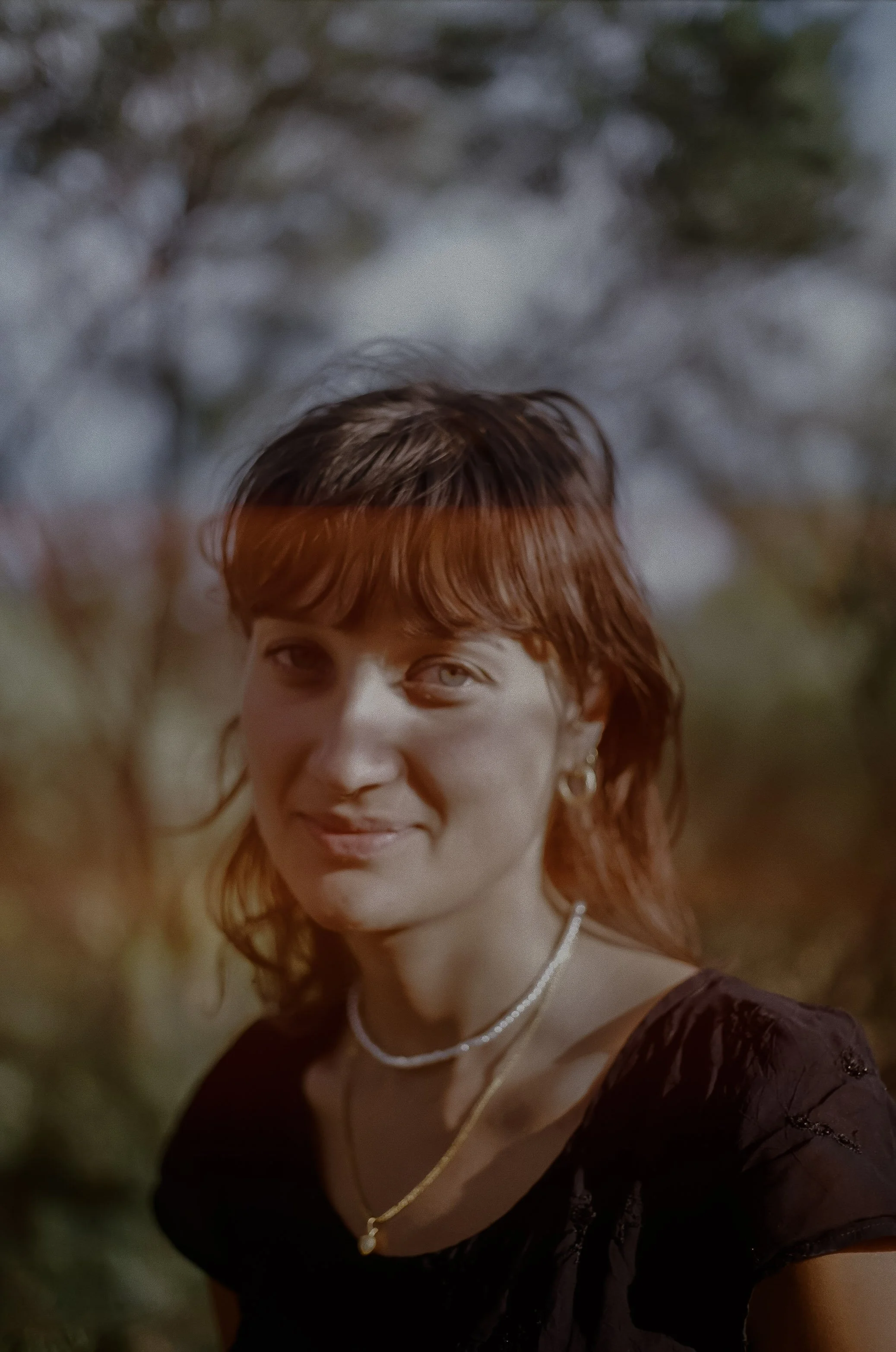 A woman with reddish brown hair smiling outdoors with trees in the background.