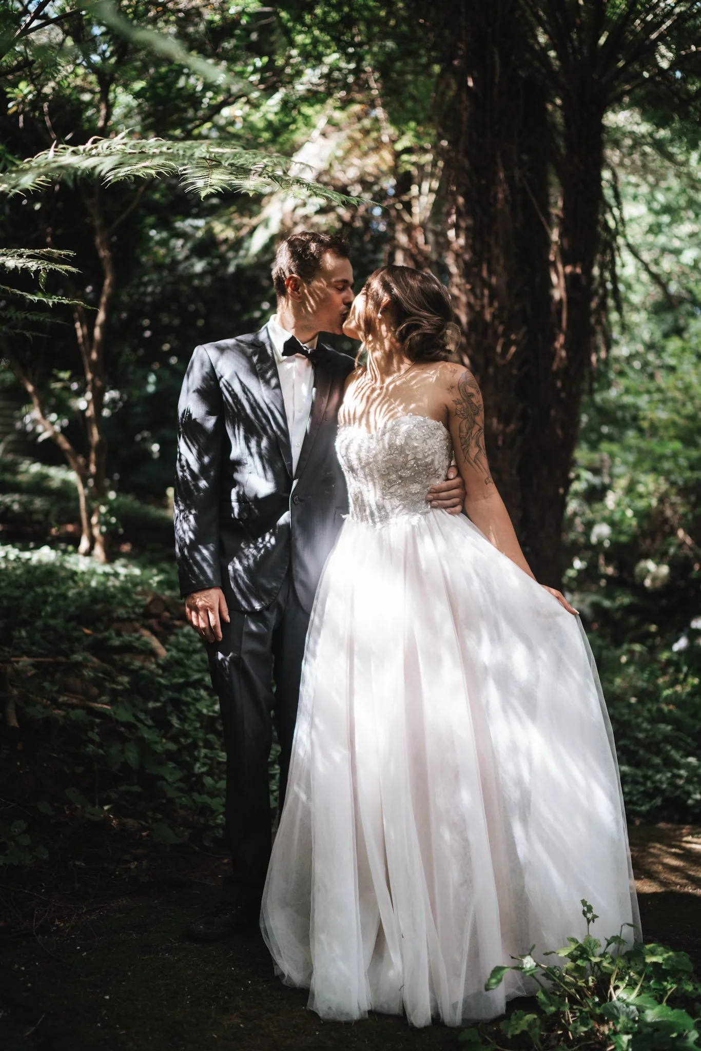 Wedding couple in a forest, kissing. The groom in a tuxedo and the bride in a white strapless wedding gown with tattoos on her arms.