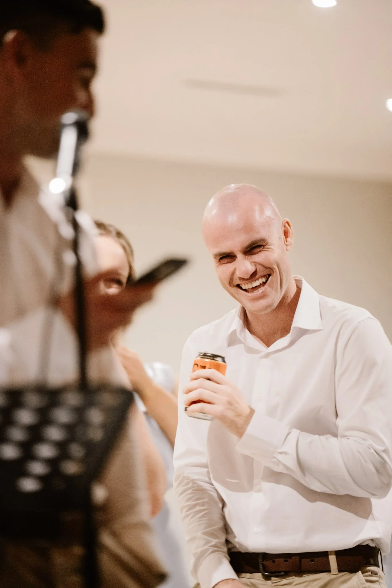 A group of people at a social gathering, with a focus on a smiling man wearing a white shirt, holding a can of beverage, and engaging with others.