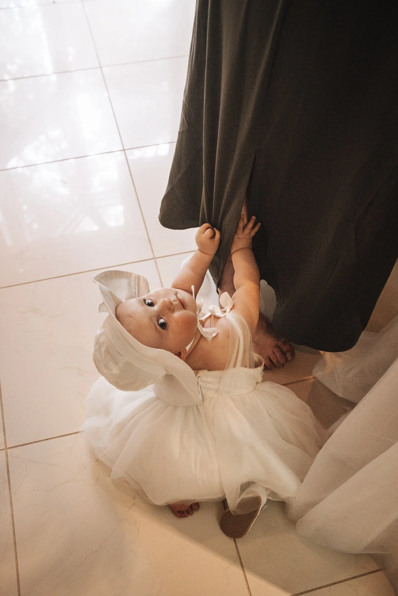 A young child dressed in a white dress and bonnet looks up at the camera while holding onto an adult's pant leg, standing on a tiled floor.