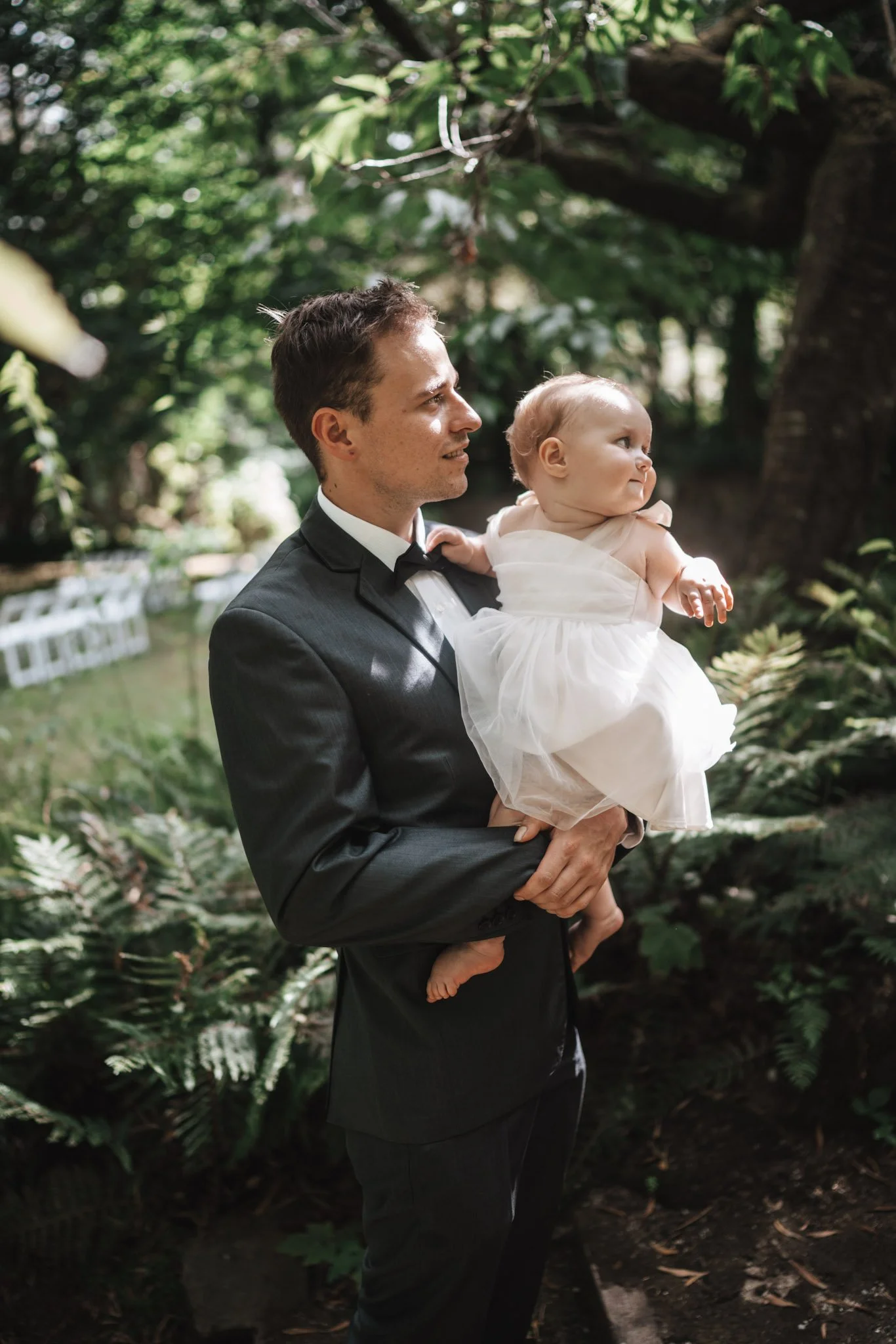 A man in a tuxedo holding a young girl in a white dress outdoors surrounded by greenery.