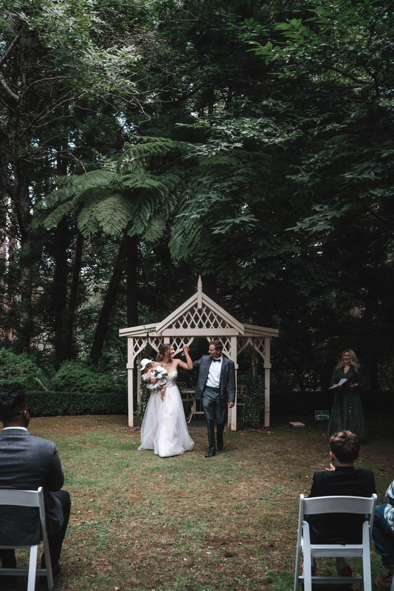 A bride and groom holding hands during their outdoor wedding ceremony, standing in front of a decorative white arbor, surrounded by lush green trees, with guests seated on white chairs.