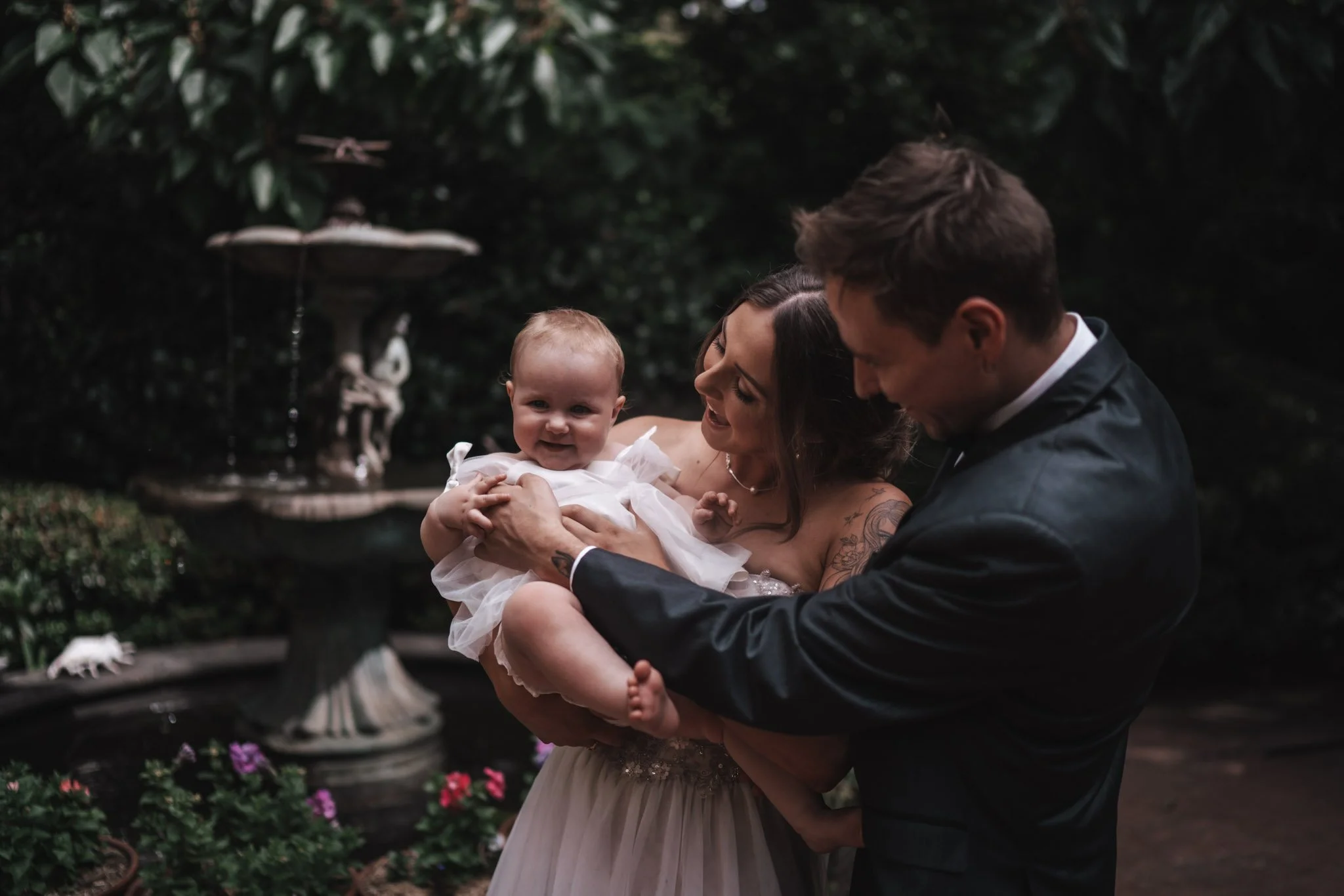 A happy family in formal attire enjoying time outdoors near a fountain with flowers, with the mother and father holding a smiling baby girl.