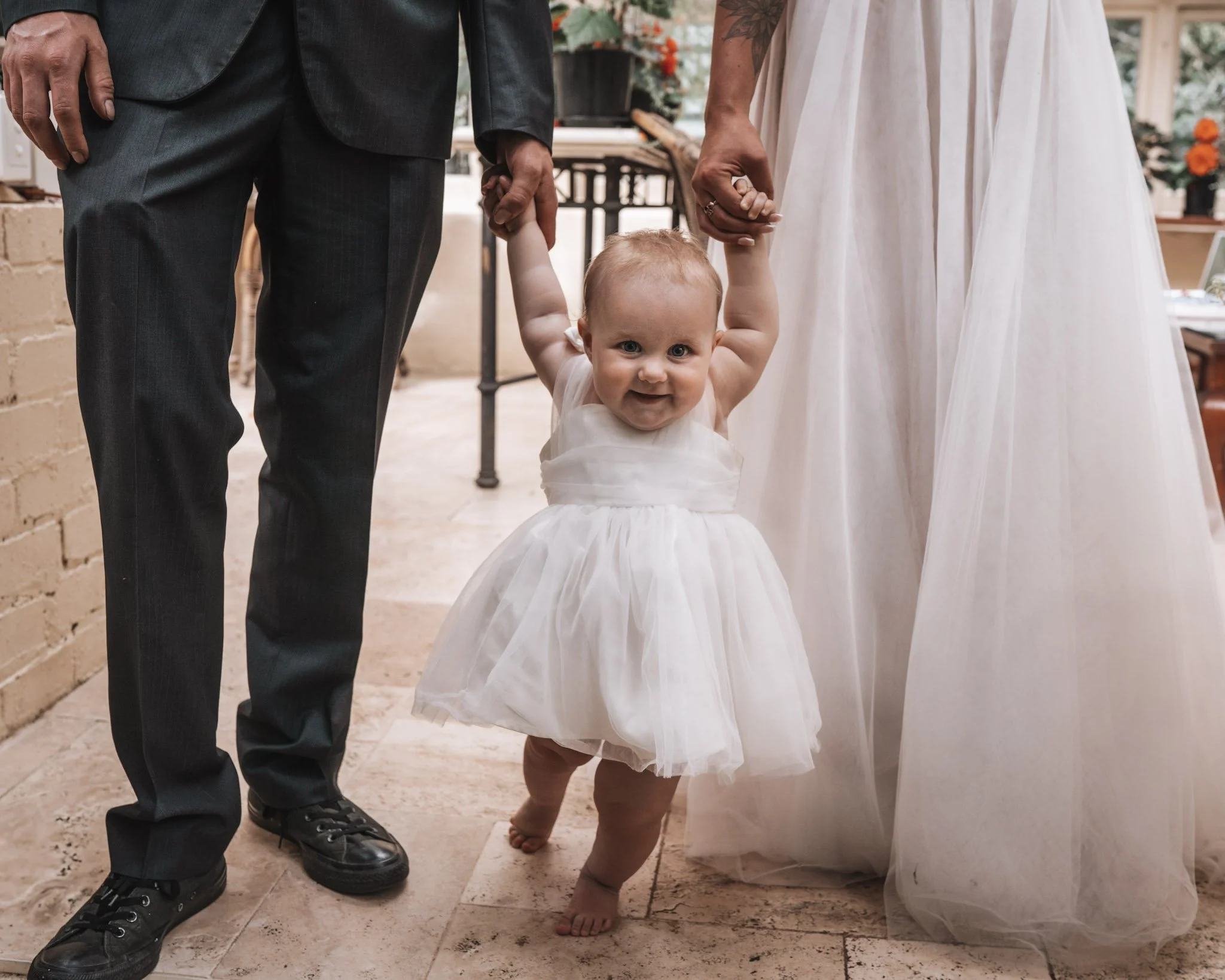 A baby girl in a white dress is holding hands with a man and a woman, walking indoors with a smile on her face.