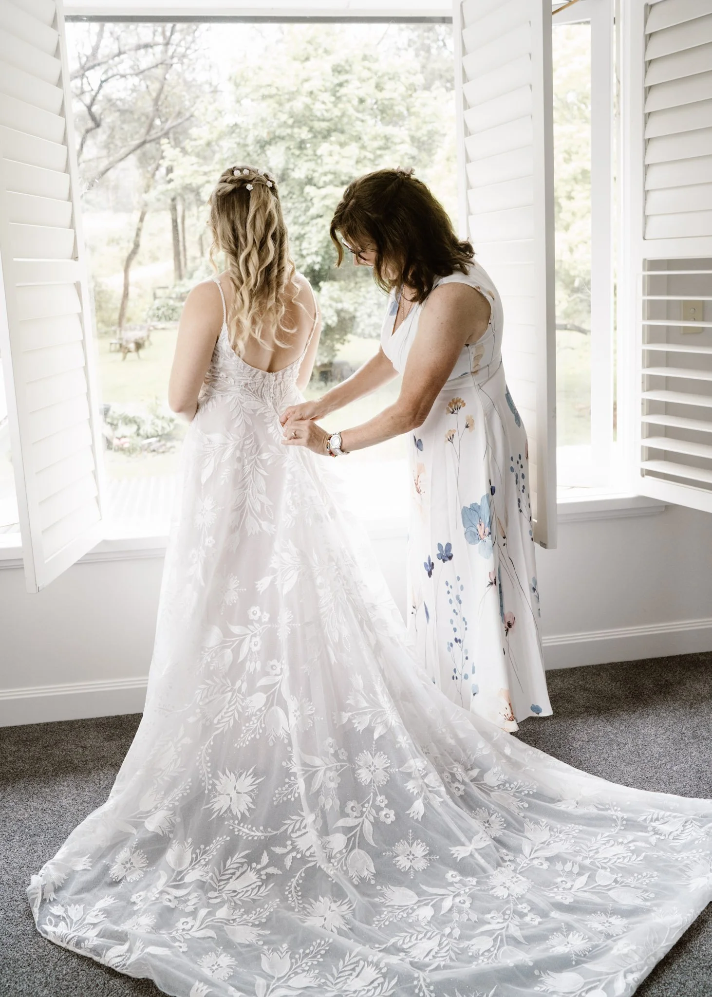 A bride getting help with her wedding dress from a woman in a floral dress, in front of open shutters with a view of a garden outside.