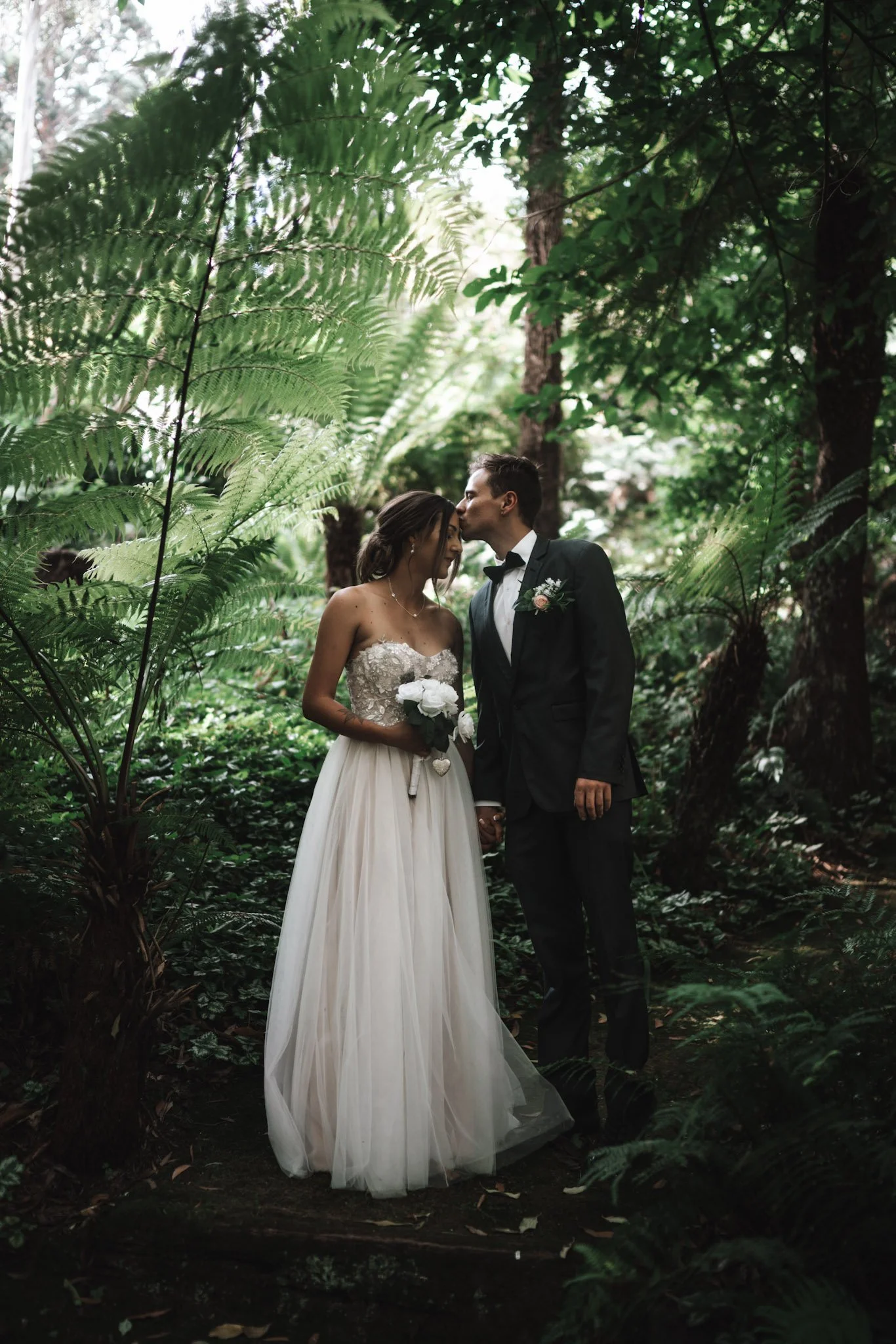 A bride and groom in wedding attire sharing a kiss in a lush, green forest setting.