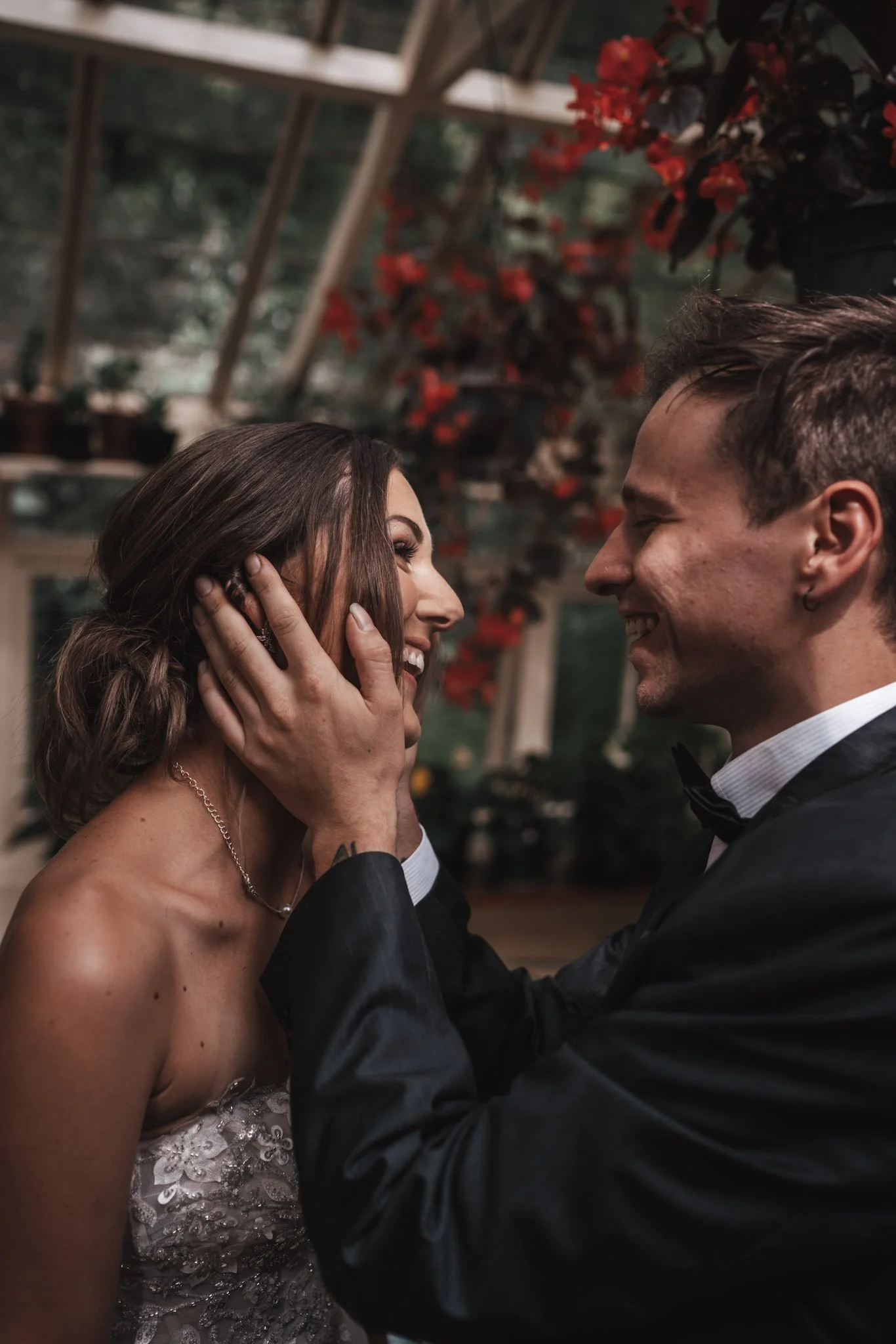 A man and woman in formal attire share an intimate moment, touching each other's faces and smiling, in a greenhouse with red flowers and greenery.