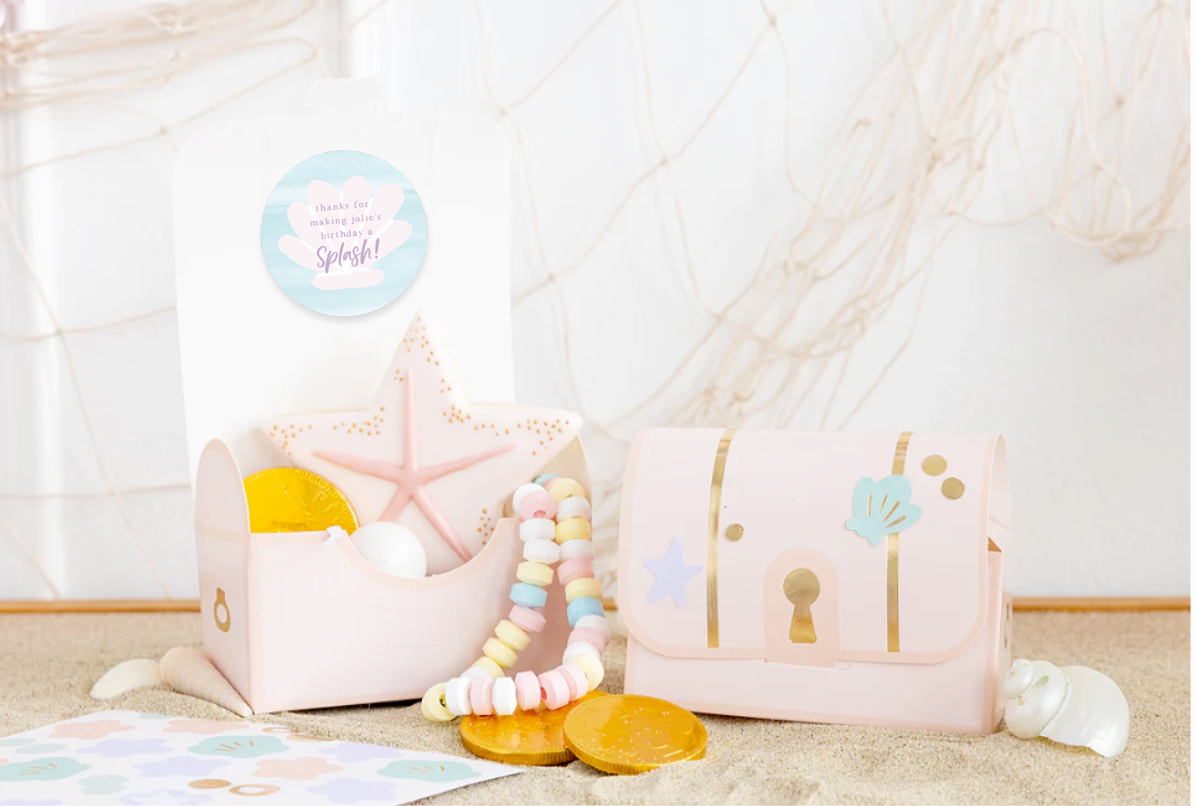 Decorative under the sea themed pink treasure chest gift box with seashells, starfish, and a string of pastel-colored beads on a beige surface with fishing net in the background.