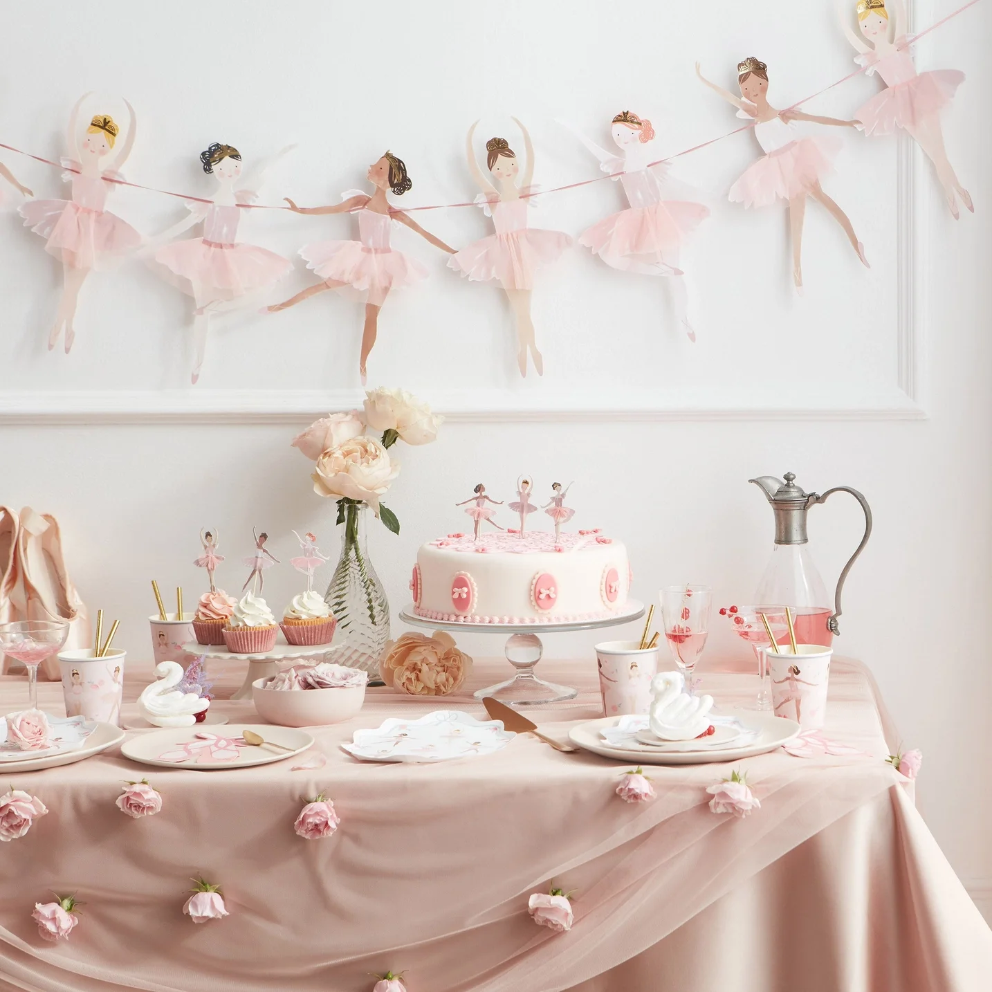 A ballet-themed party table decorated with pink and white items, including a banner of paper ballerinas, a pink cake with ballerina toppers, cupcakes, and floral accents.