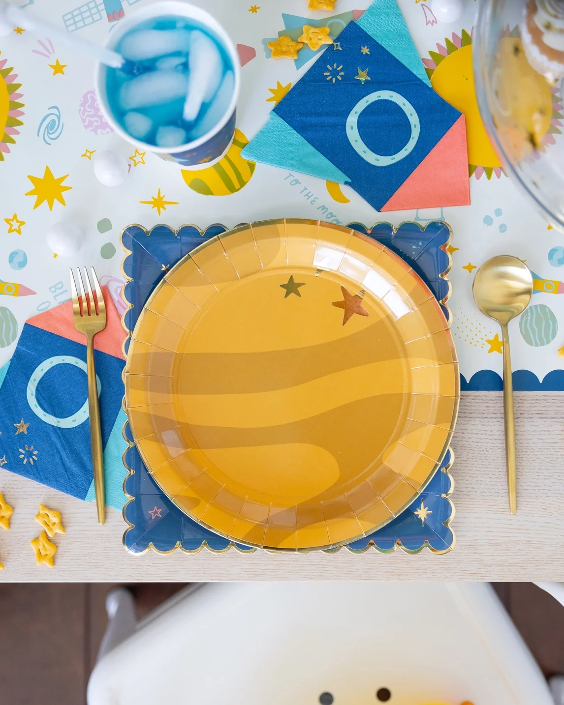 A colorful birthday table setting with a yellow plate, gold fork and spoon, blue and pink napkins, a partly visible drink with ice, and star-shaped decorations.