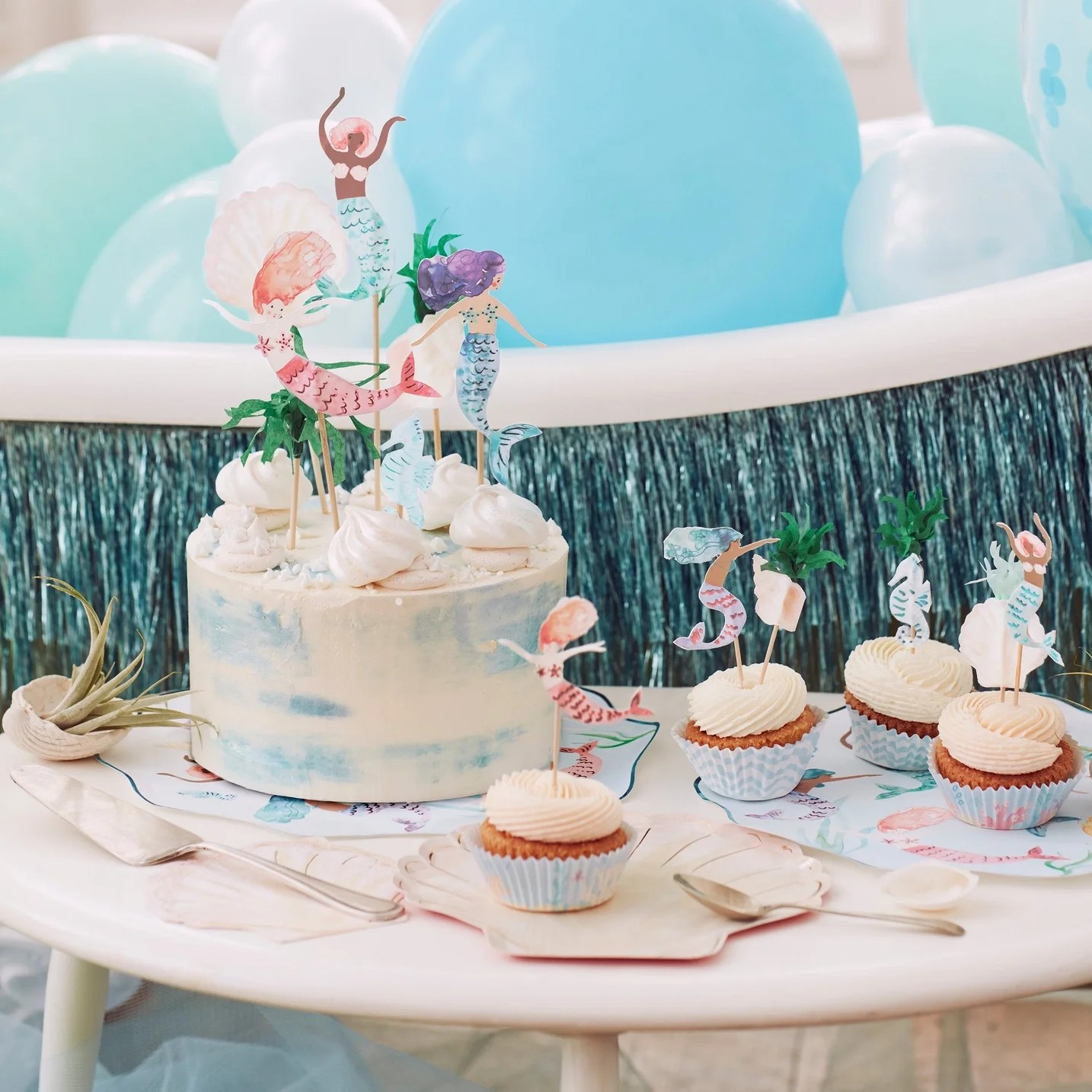 A birthday cake decorated with ocean-themed mermaid toppers and cupcakes with mermaid toppers, surrounded by pastel balloons and ocean colors.