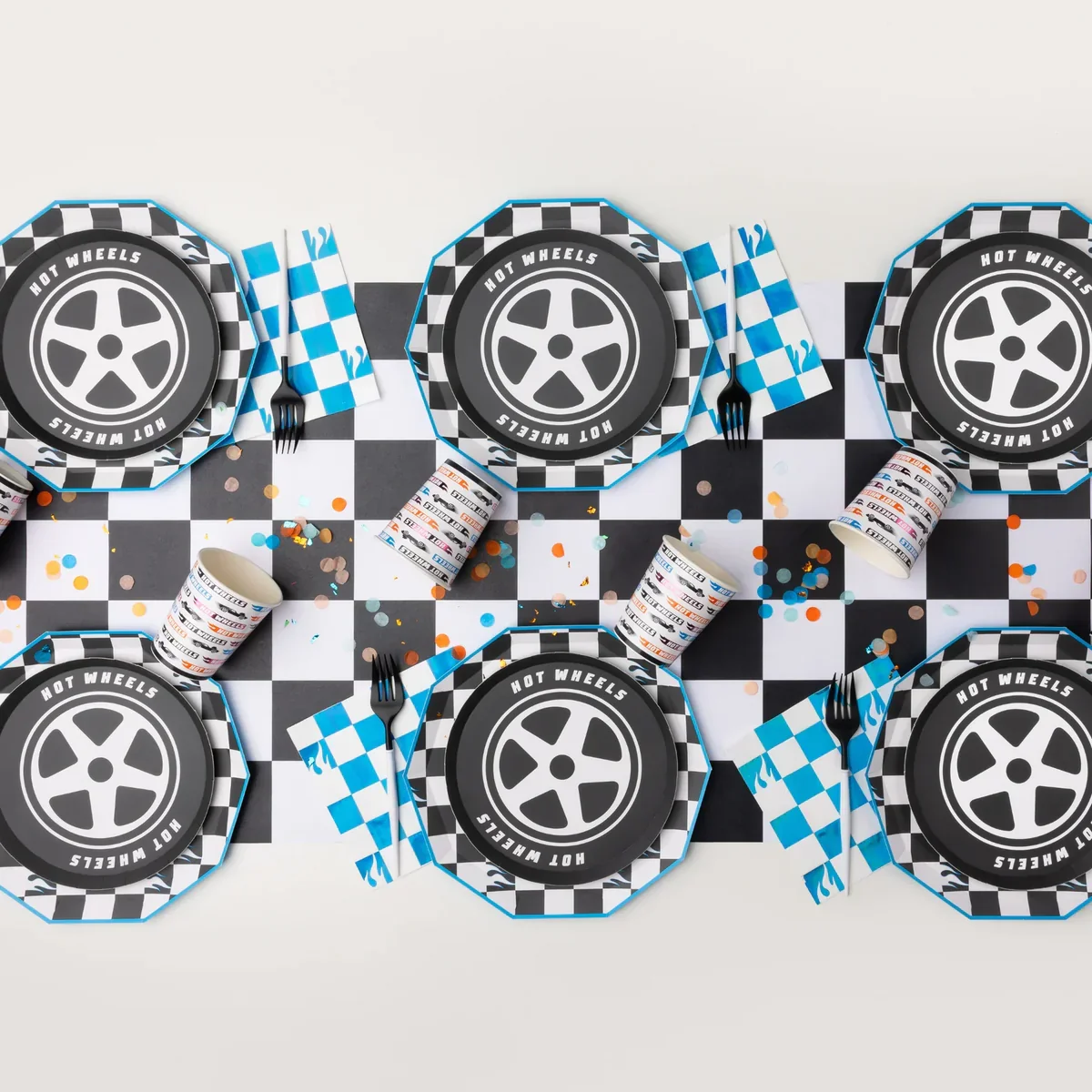 Race car-themed party table setting with checkered black and white tablecloth, black plates featuring a car wheel design and the words 'Hot Wheels' and 'Hot Wheels' around the rim, blue checkered napkins, black forks, white cups with racing-related text, and scattered colorful confetti.