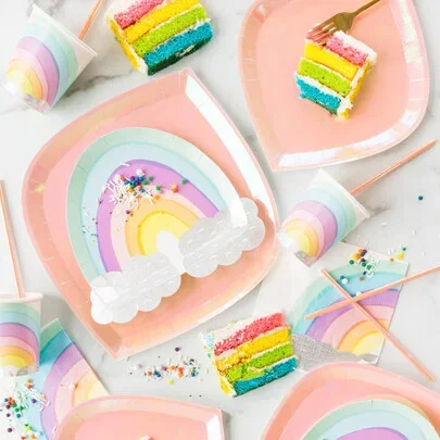 Colorful rainbow cake slices with rainbow-themed party supplies on pink plates and star-shaped balloons.