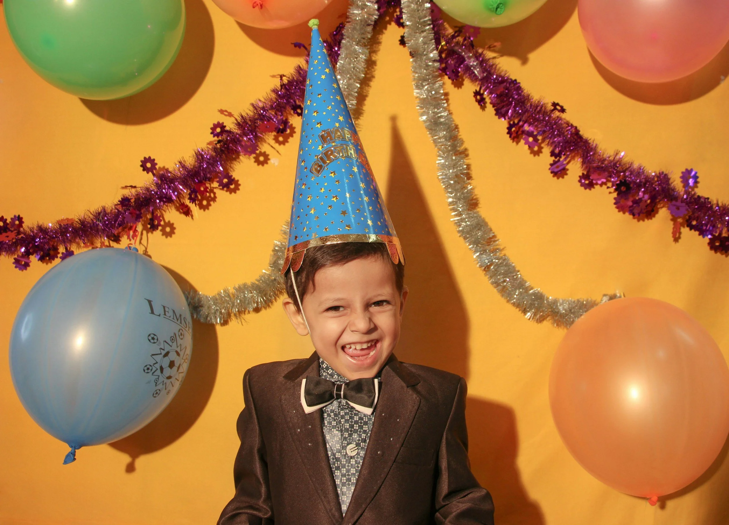 A young boy at a birthday party wearing a blue birthday hat with gold stars and 'Happy Birthday' written on it. He is smiling and dressed in a brown suit with a bow tie. The background features colorful balloons and purple and gold tinsel decorations on a yellow wall.