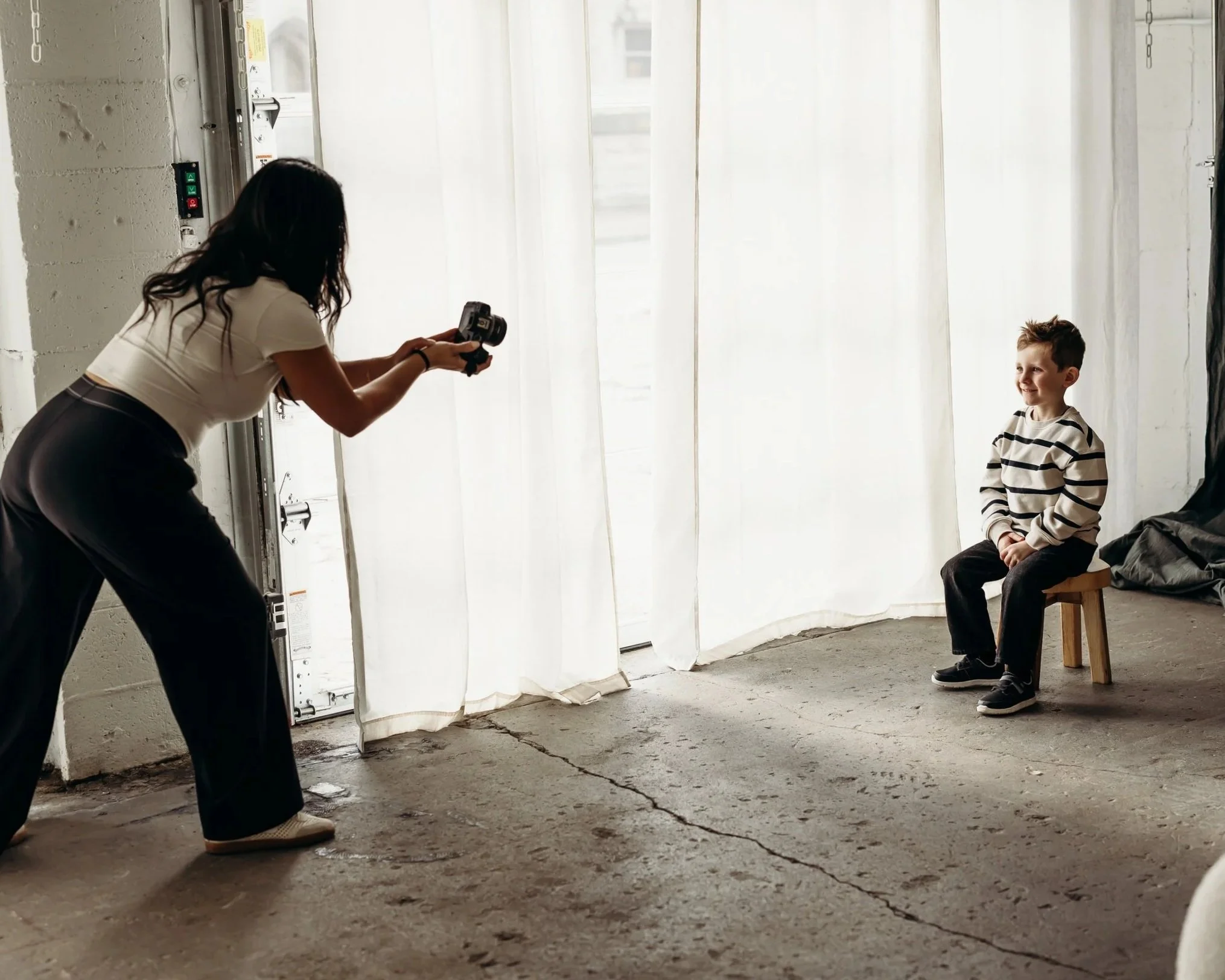 A woman takes a photo of a young boy sitting on a stool in front of a white curtain backdrop, with studio lighting and concrete floor in a photography studio.