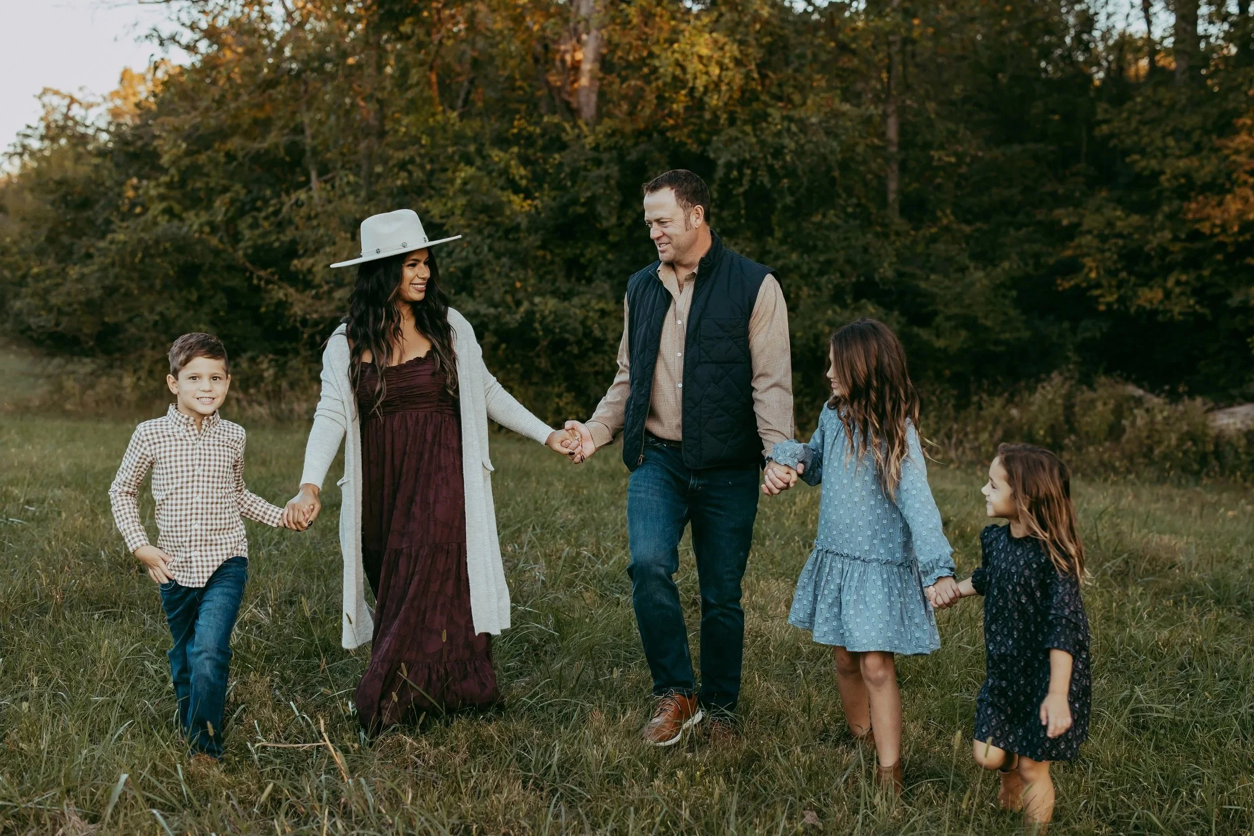 A family of six holding hands and walking across a grassy field with trees and orange autumn leaves in the background. The woman is wearing a large light-colored hat and a long dress with a cardigan, while the man is wearing a vest over a button-up shirt. They are smiling and enjoying the outdoor moment.