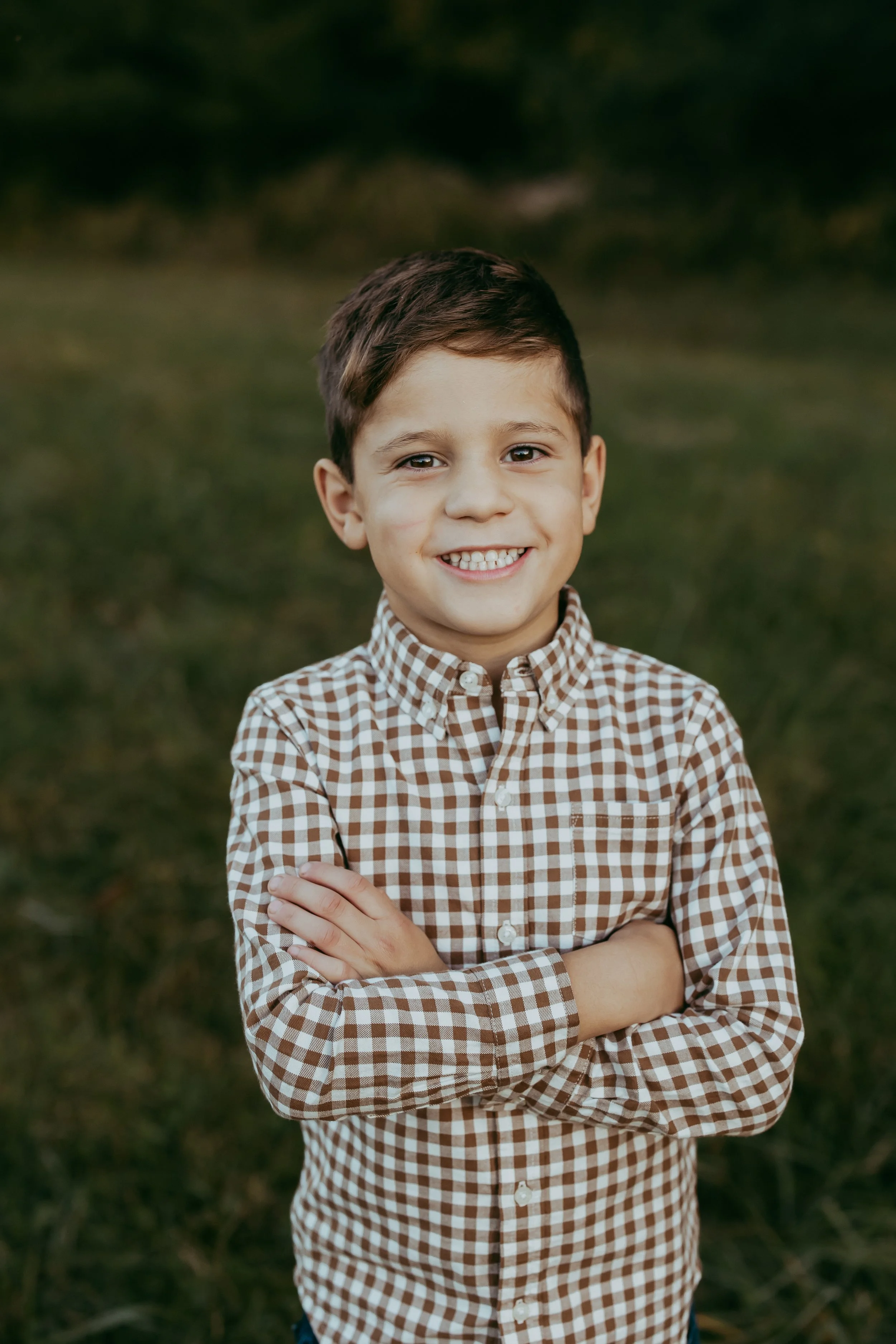A young boy with dark hair, smiling, standing outdoors on grass with arms crossed, wearing a brown and white checkered shirt.