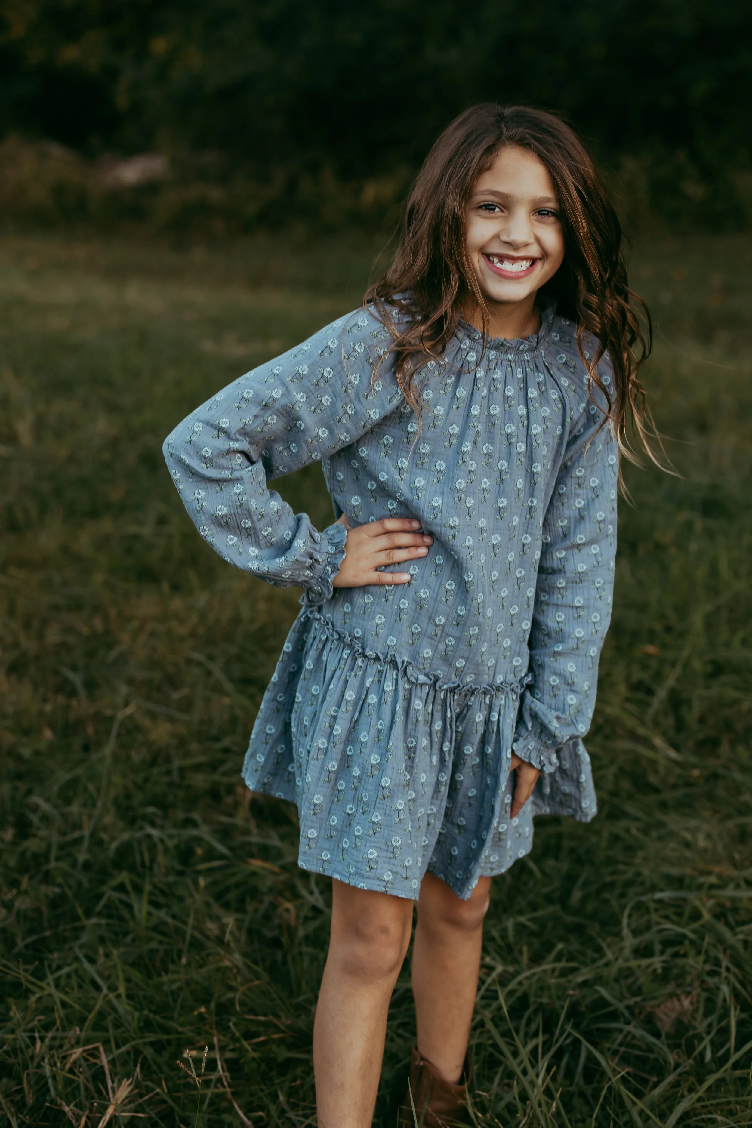 A young girl with long, wavy brown hair, smiling with her teeth showing. She is wearing a blue long-sleeve dress with a floral pattern and ruffled hem, standing outdoors in a grassy field with trees in the background.