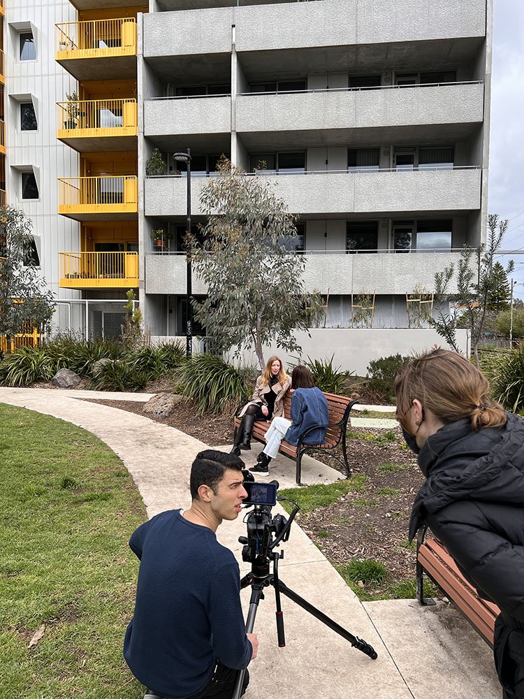 Julie interviews Rebecca sitting on a park bench, in front of an apartment building with yellow balconies.