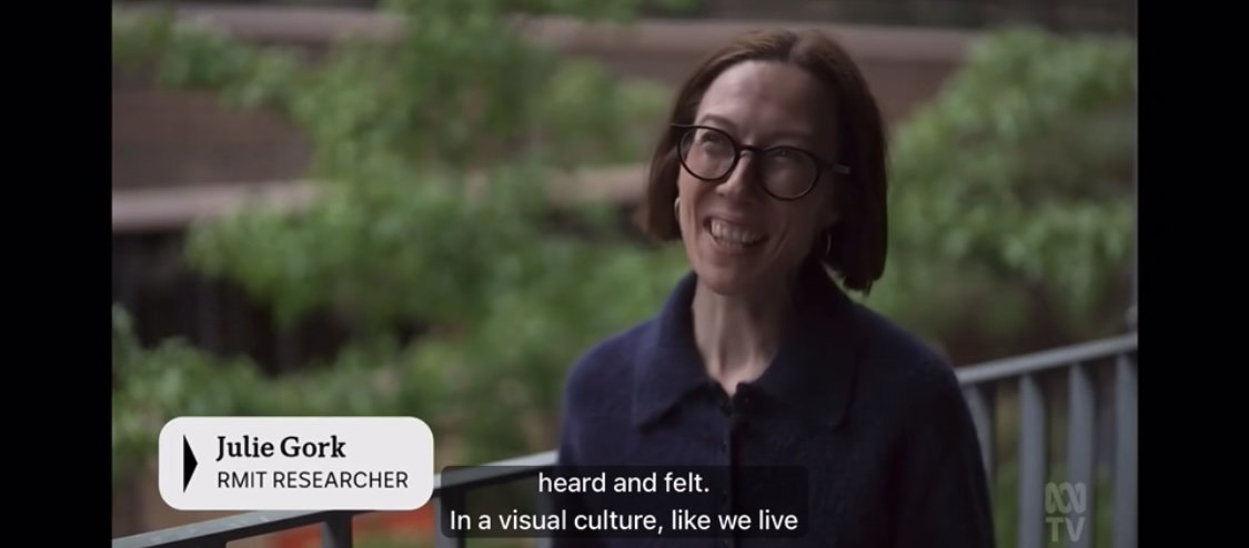 A screen shot from the ABC news of Julie giving an interview. She has short brown hair, glasses and wears a navy blue cardigan.