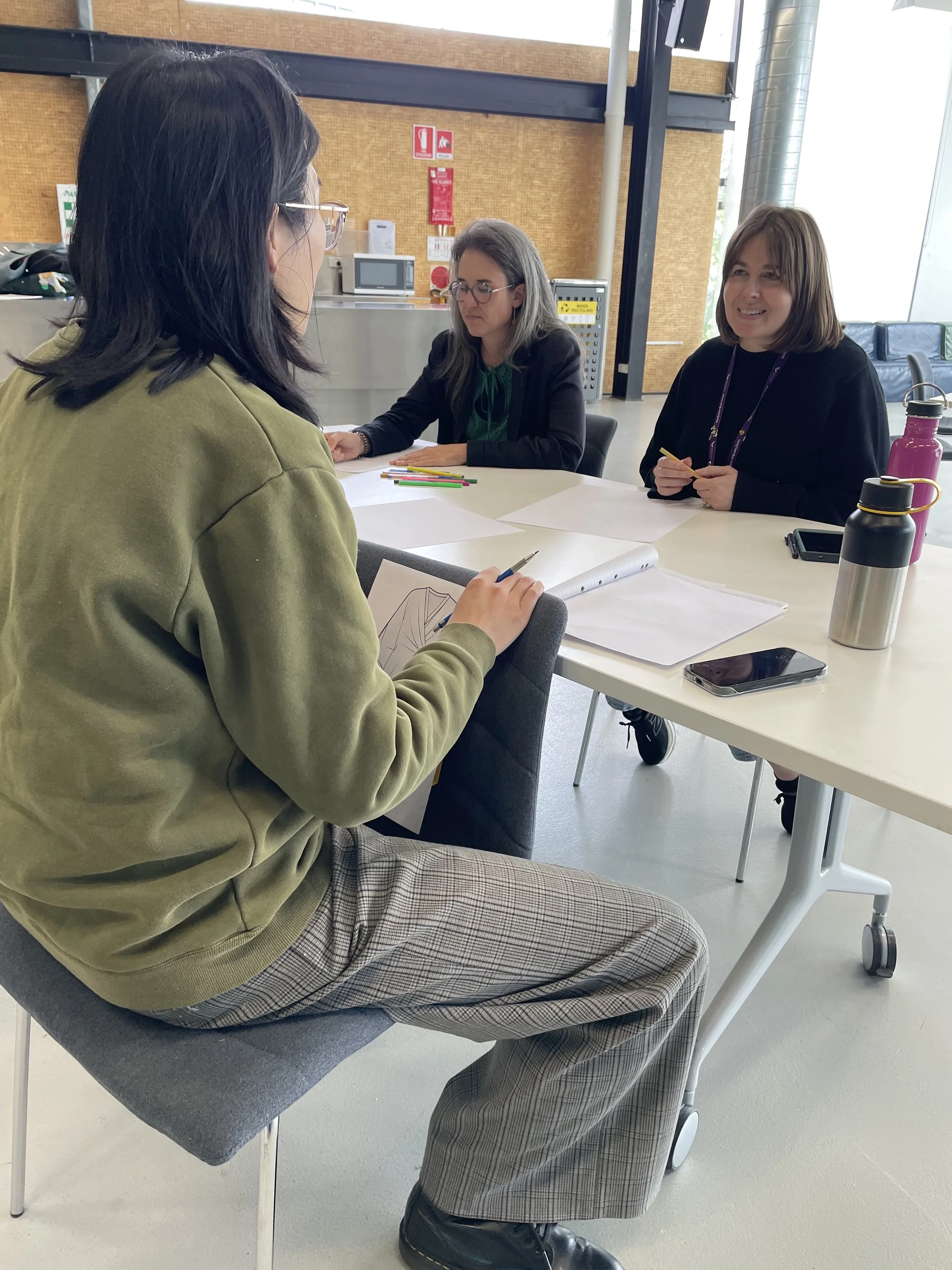 Three women engaged in a discussion or interview, seated at a white table with notebooks, pens, and water bottles in an indoor space with industrial decor.