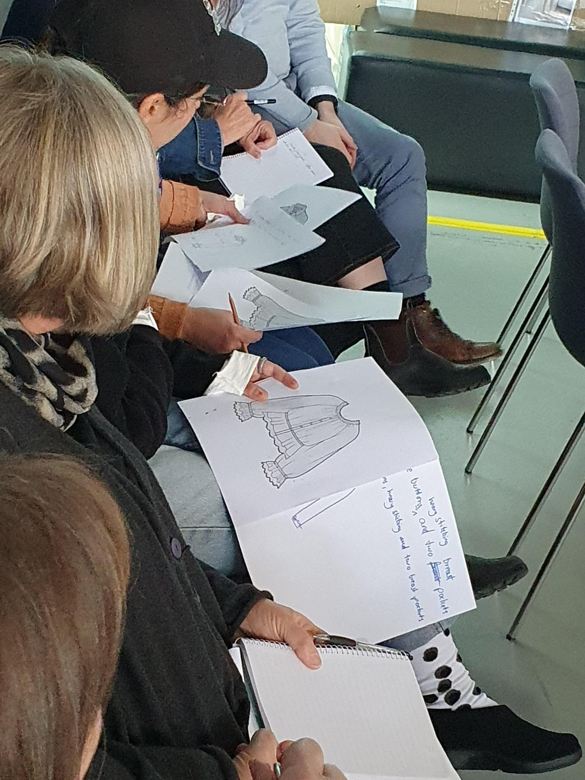 People seated in a row, drawing and coloring pictures of dresses and clothing on sheets of paper, in a classroom or workshop setting.