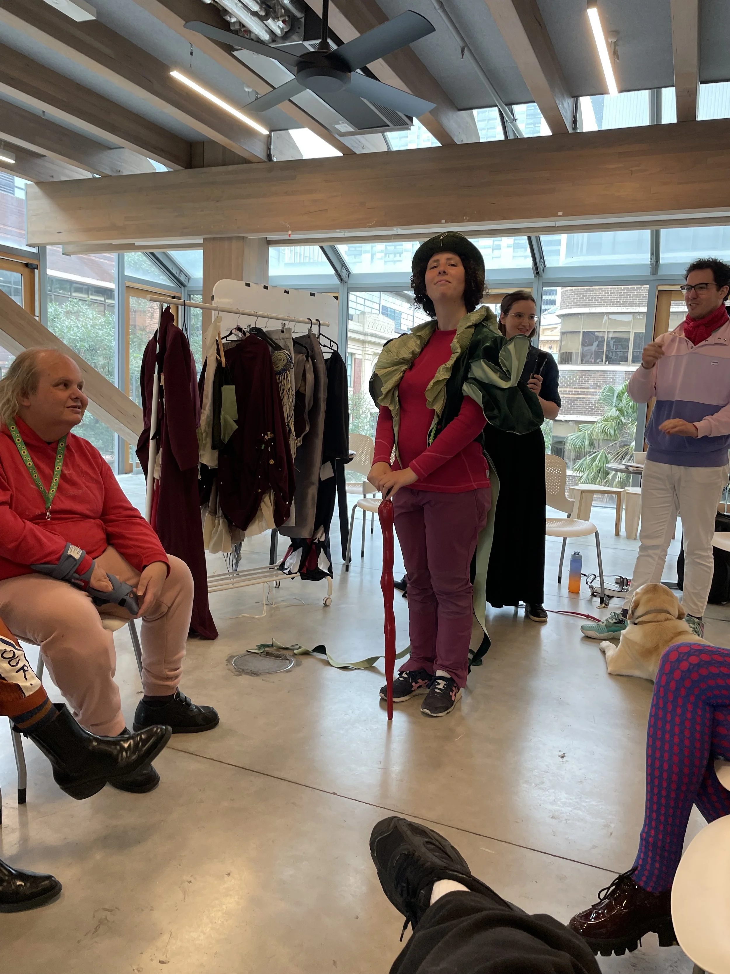 A woman wearing a green capelet, bowler hat, pink top and purple pants proudly holds her head high while starting to walk down the runway. She holds an elongated capsicum walking stick, creating an Alice in Wonderland effect.