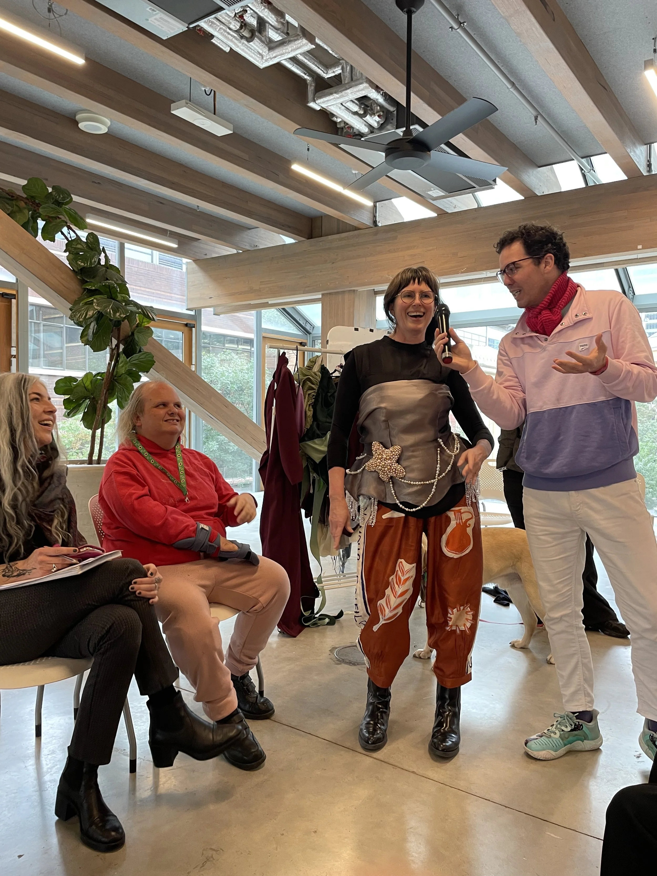 A woman stands at the end of a make-shift catwalk wearing a corset top and baggy pants. A man stands next to her attempting to describe her outfit as she walks down the runway.