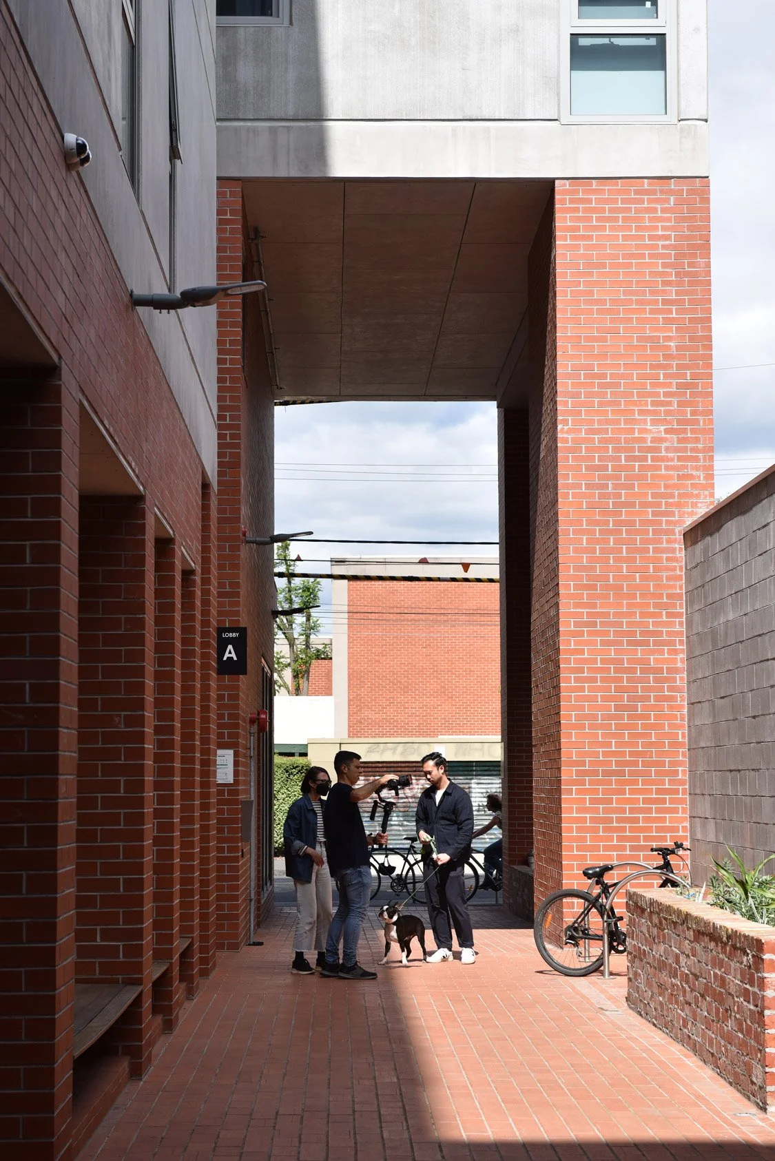 Julie, a videographer, and a man with dog are shooting a film in walkway by a red brick apartment building.
