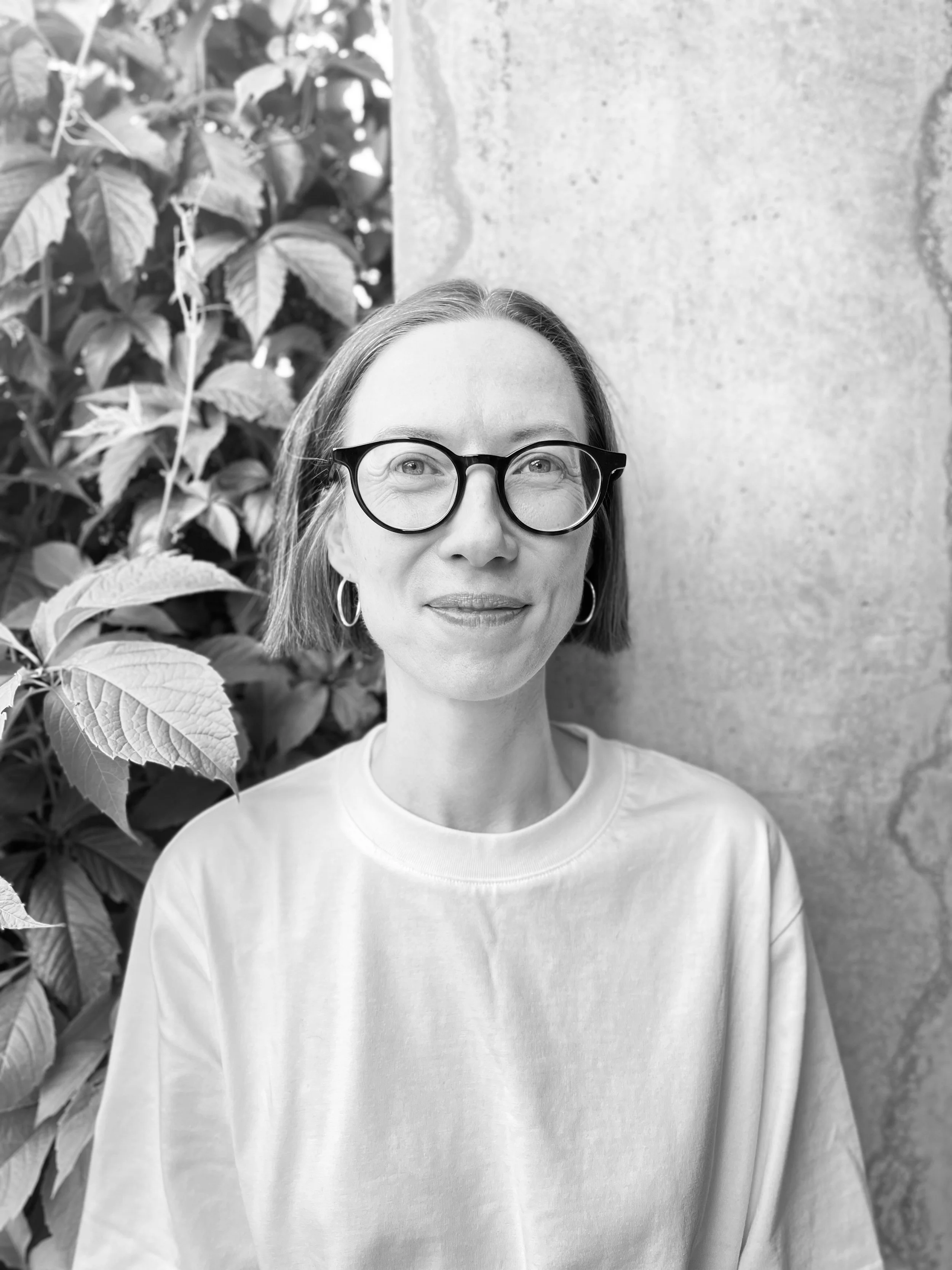 A woman with glasses and hoop earrings standing outdoors with foliage on her left and a textured wall behind her.