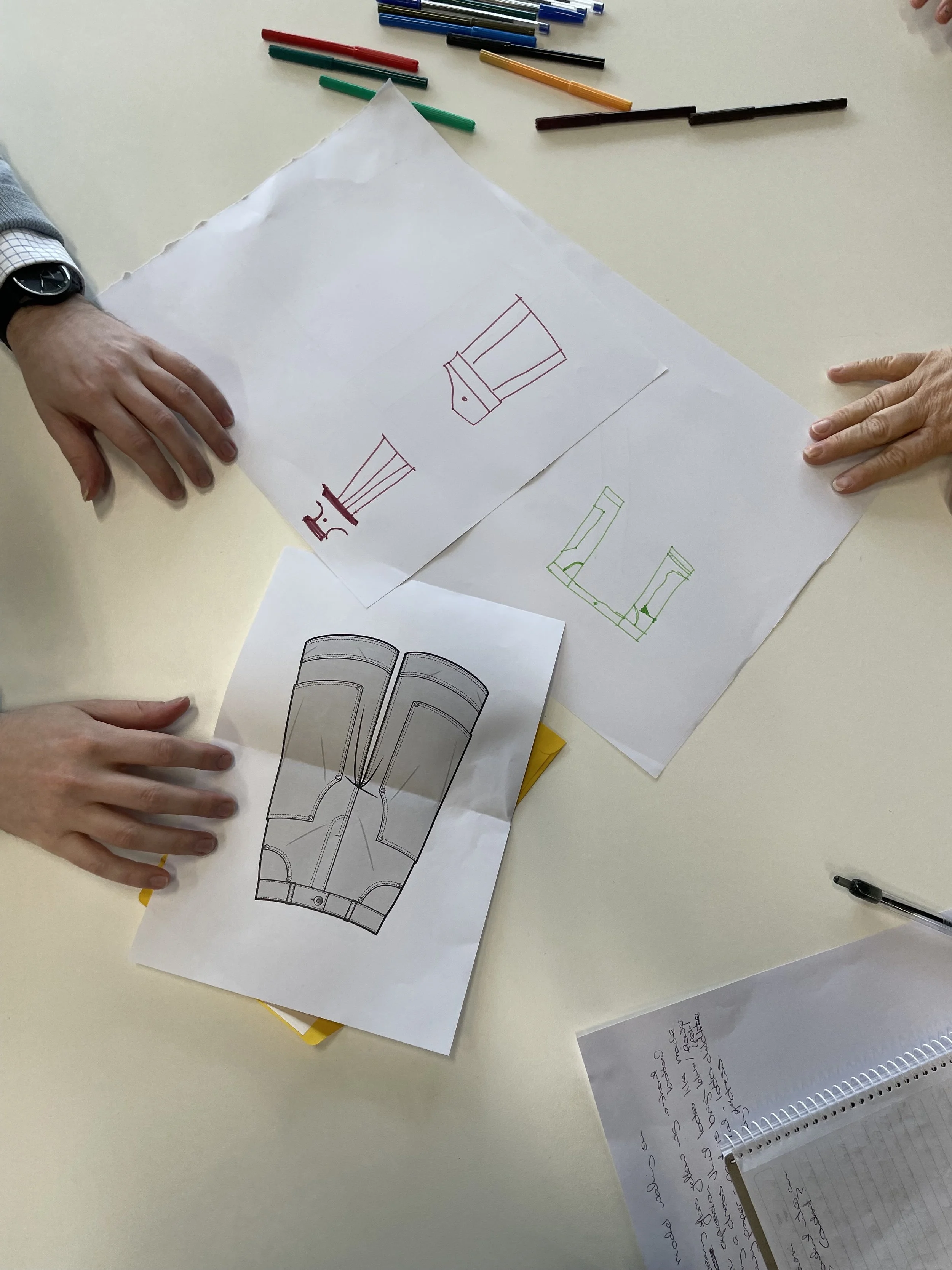 Two people examining technical drawings and sketches of footwear designs on a table, with colored markers and notes.