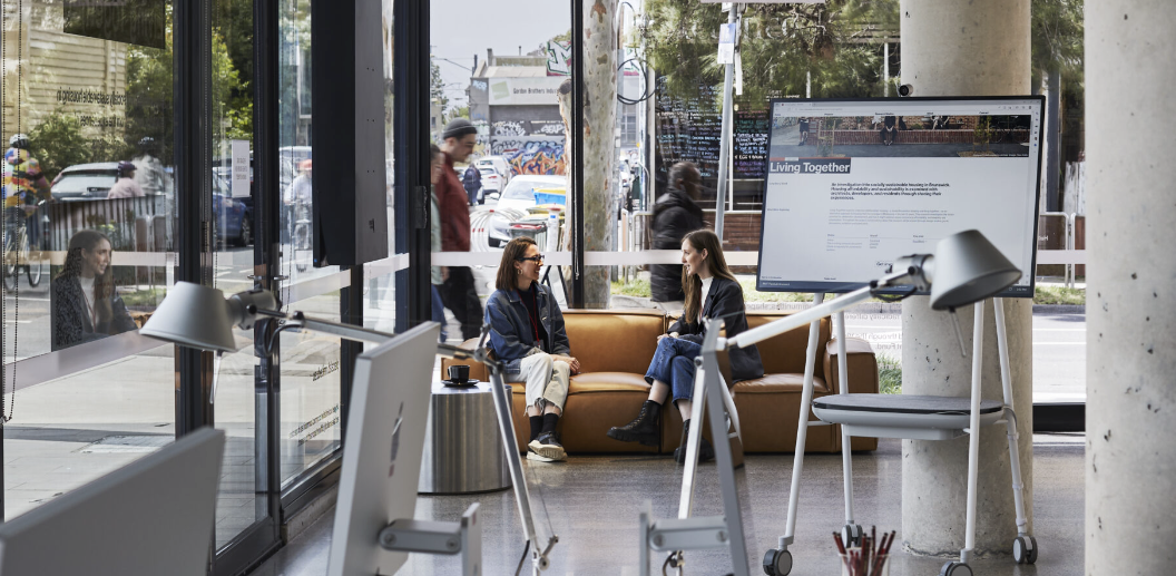 PlaceLab Researchers, Julie and Hayley, are having a conversation on a brown leather couch in a the light-filled urban research studio.