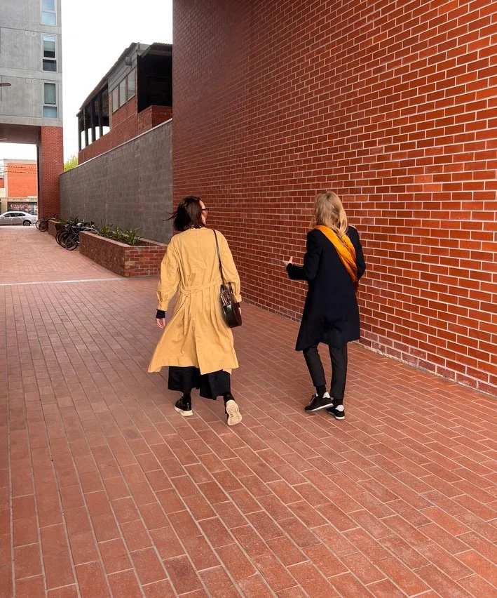 Julie, wearing a yellow trench, walk with Rebecca, dressed in black with orange scarf.