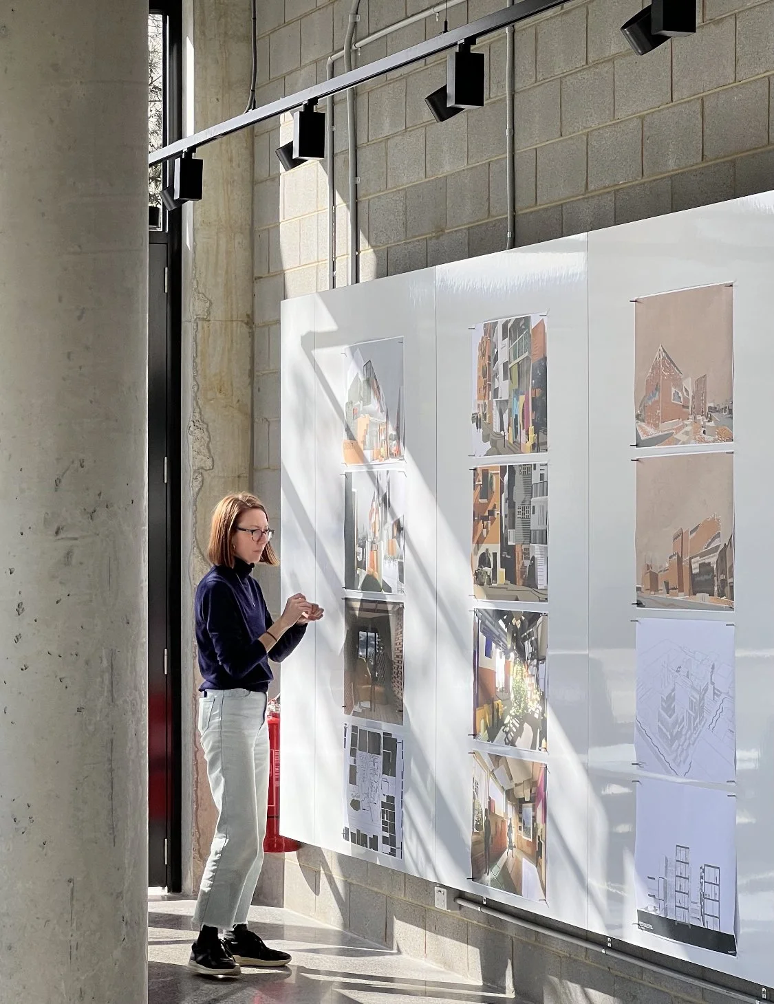 Julie, wearing light denim jeans and a navy turtleneck, is pinning architectural drawings on a wall for an exhibition.