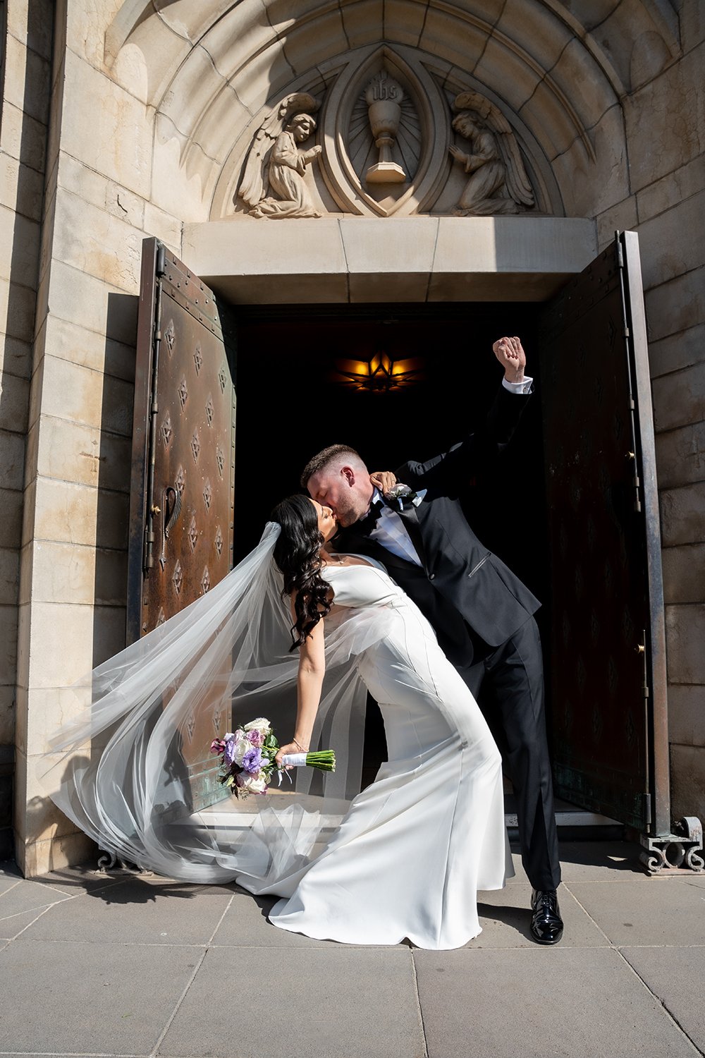 A bride and groom kissing in front of a church doorway, with the groom lifting the bride. The bride is in a white wedding dress with a flowing veil and holding a bouquet of purple and white flowers. The brick arch and religious sculptures are visible