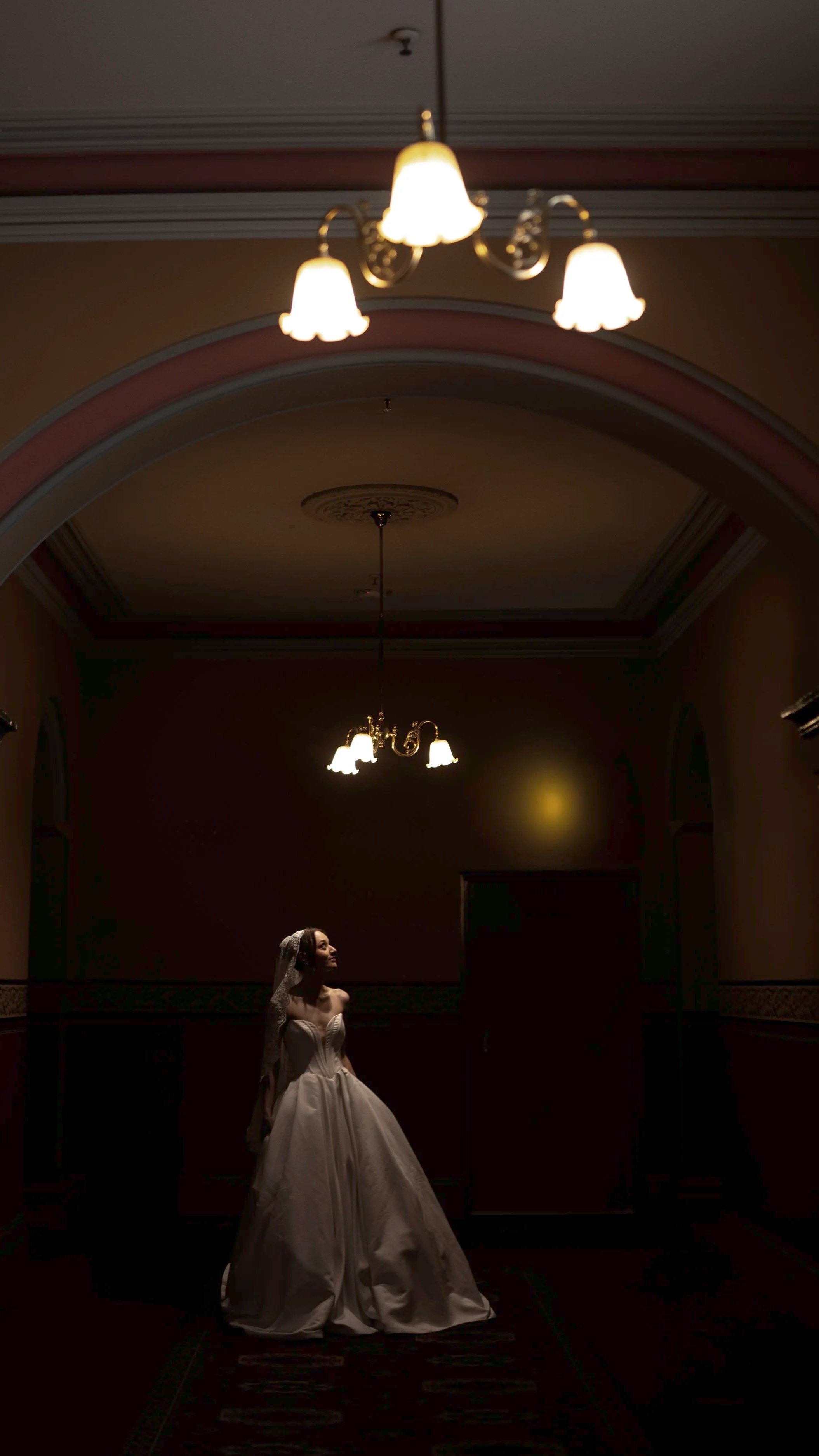 A woman in a wedding dress standing in a dimly lit room with ceiling chandeliers.