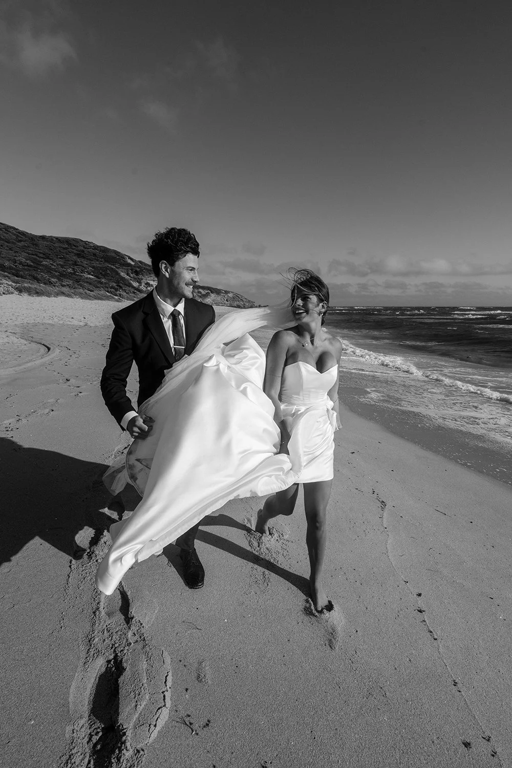Black and white photo of a couple in wedding attire walking barefoot along the beach, smiling and laughing with the ocean in the background.