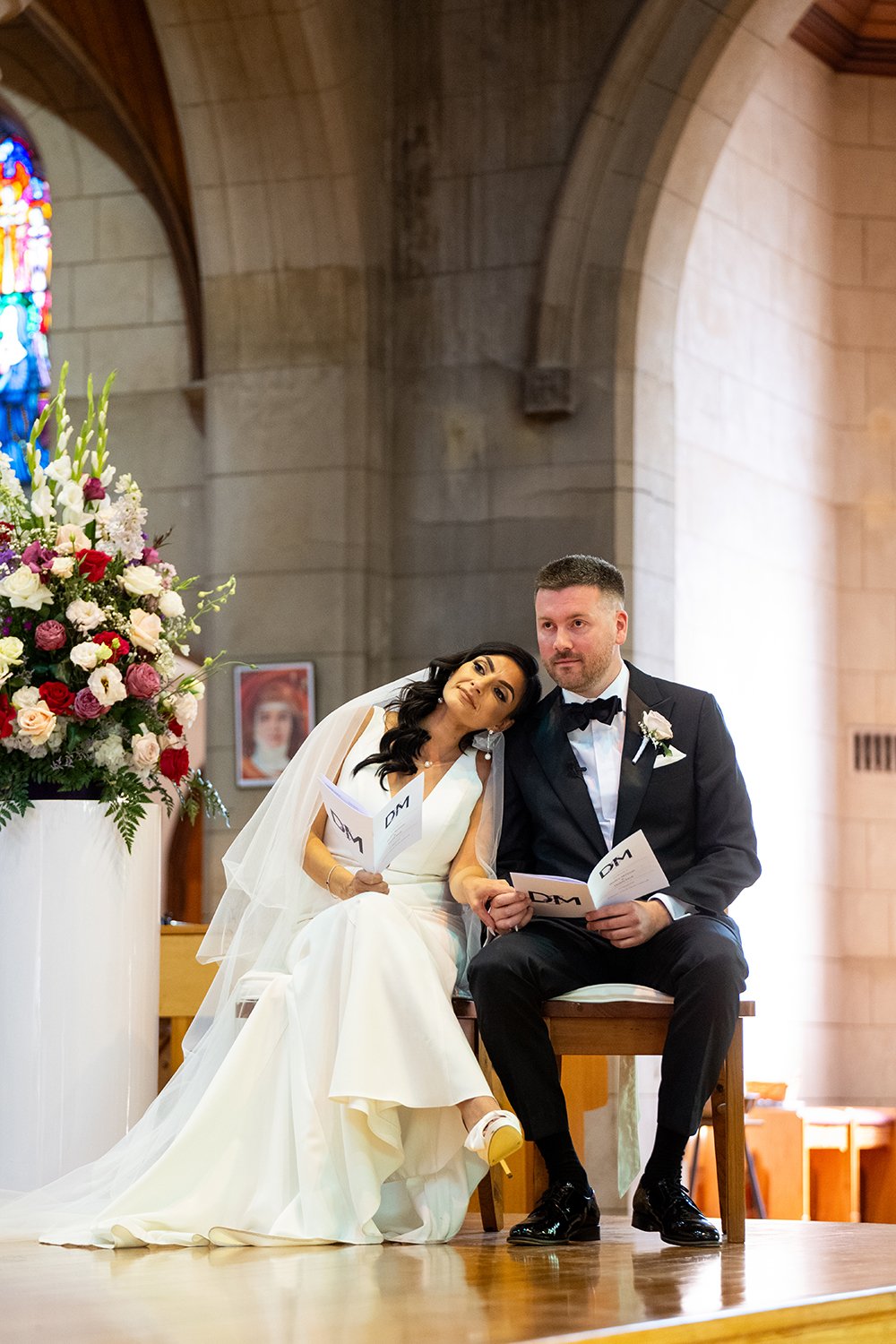 A bride and groom sitting together on a bench in a church during their wedding ceremony. The bride is leaning her head on the groom's shoulder, and both are holding wedding programs. The church has high arched ceilings and a large flower arrangement nearby.