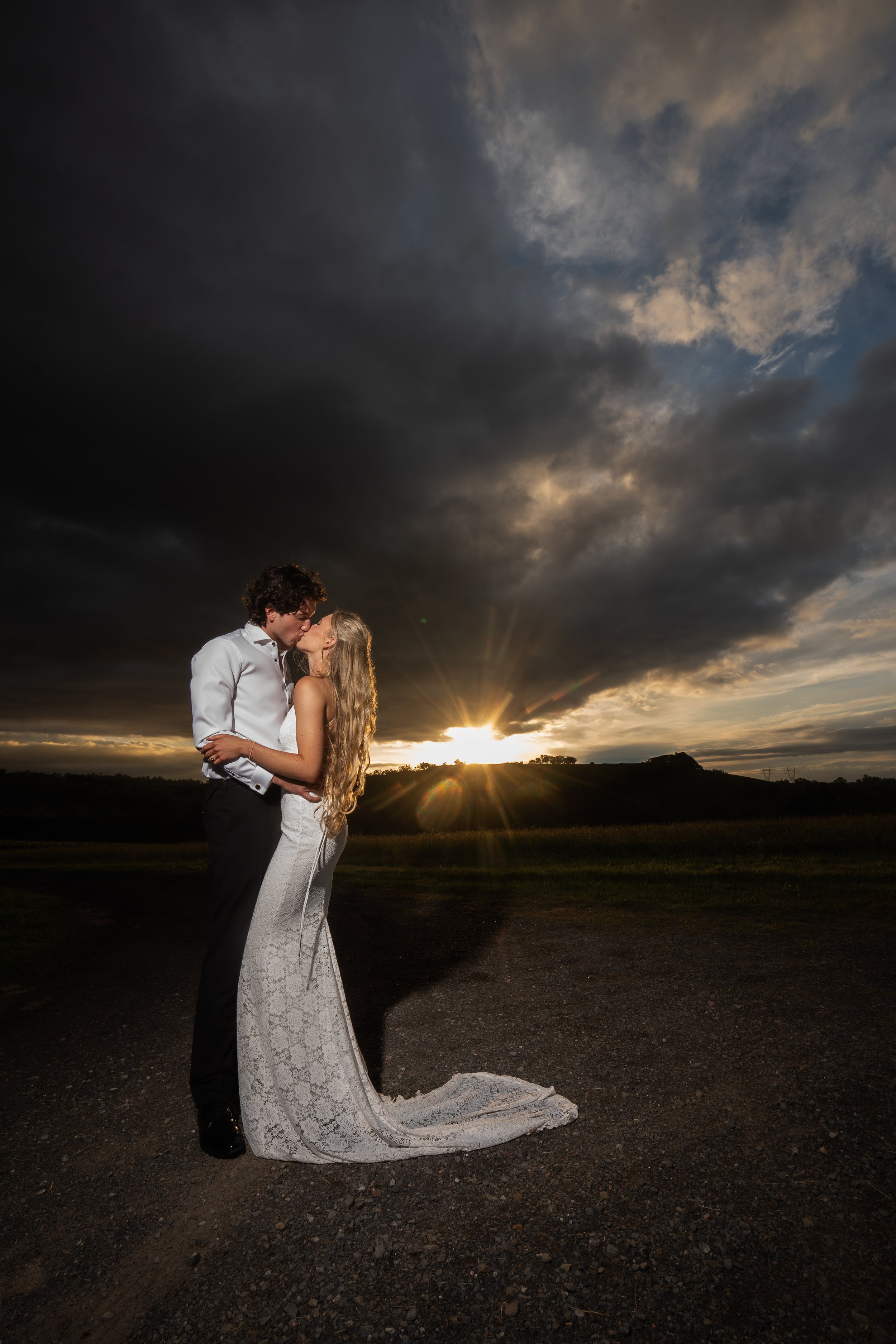 A couple in wedding attire sharing a kiss outdoors at sunset with dark clouds overhead and a scenic landscape