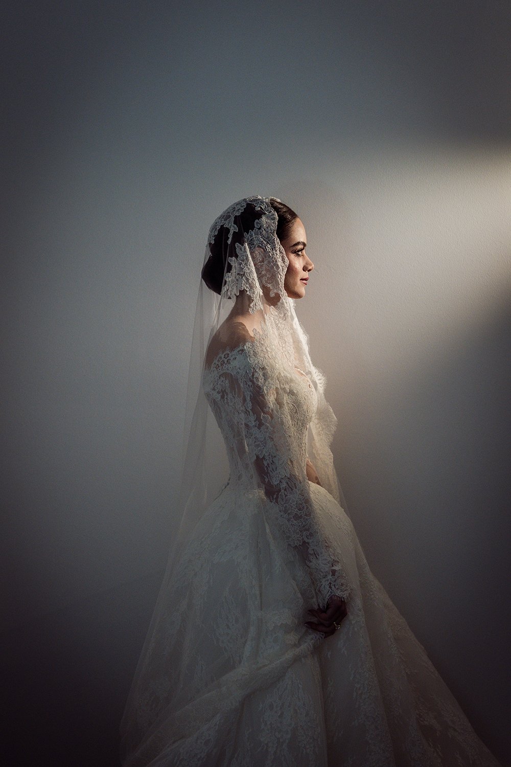 Profile of a woman in a wedding dress and lace veil standing against a plain wall.