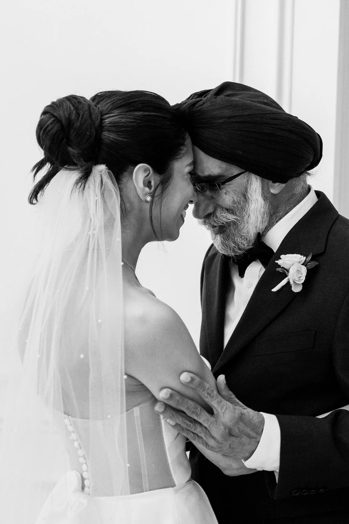 A bride and her father, share a tender moment with their foreheads touching. The bride is wearing a strapless wedding gown and a veil, while the man is dressed in a tuxedo with a boutonnière and a turban.
