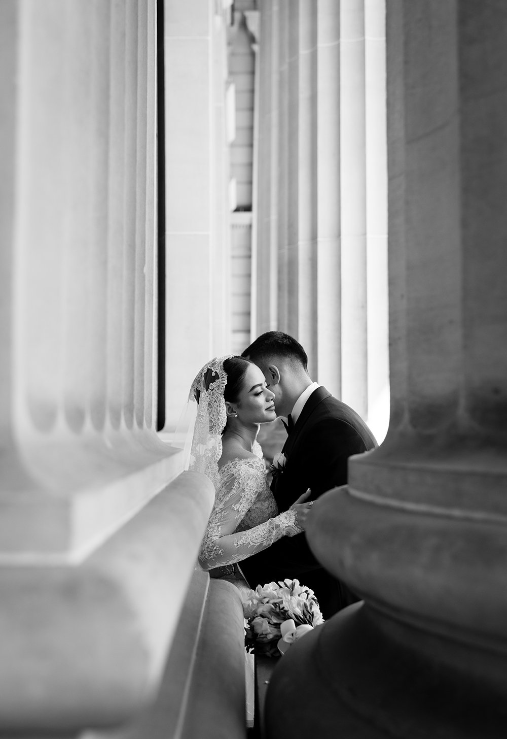 A bride and groom embrace in a black-and-white photo, standing between large classical columns, with the bride wearing a lace wedding dress and veil, and the groom in a tuxedo holding a bouquet of flowers.