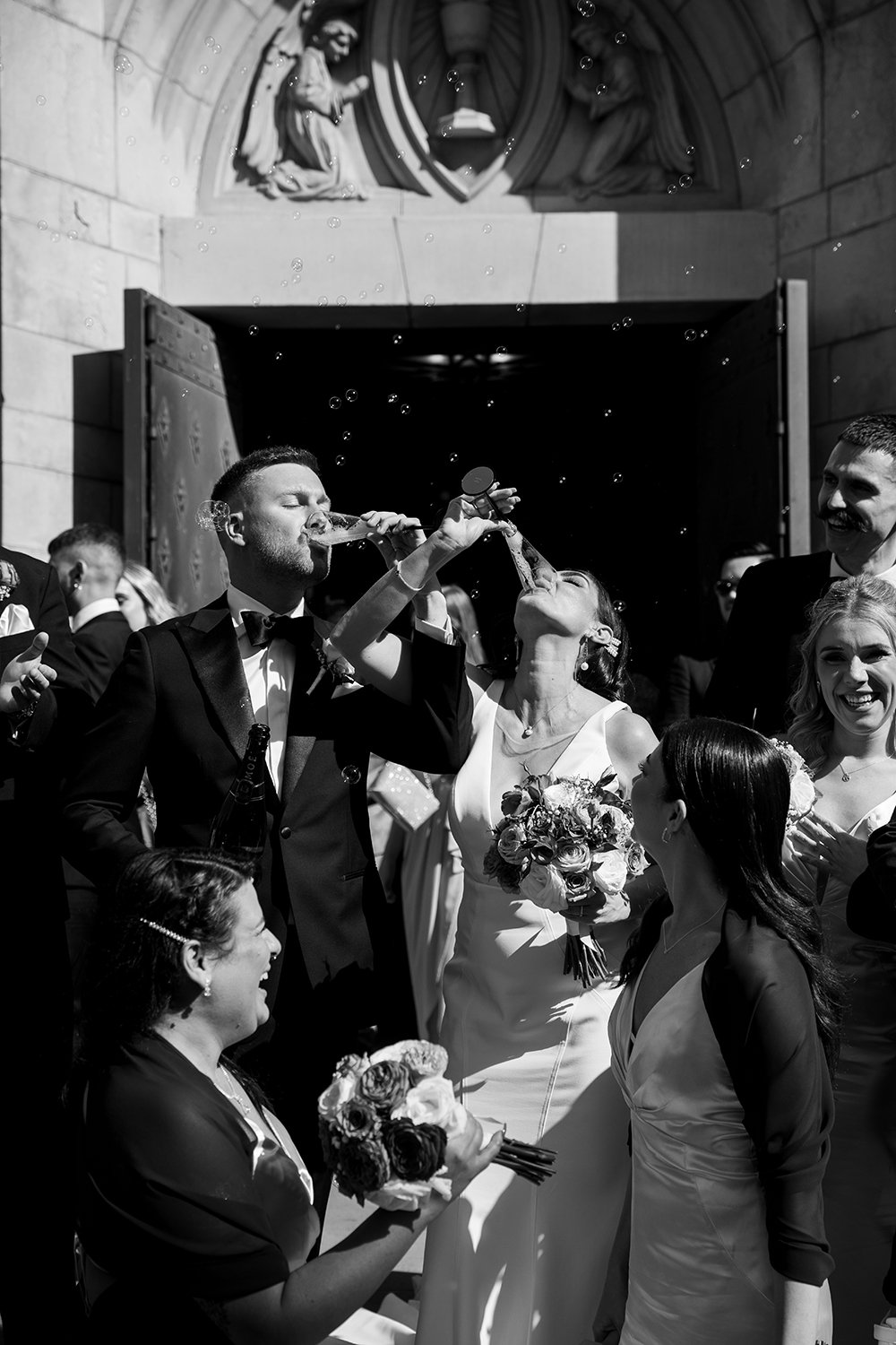 Black and white photo of a bridal couple leaving church with people dressed in formal attire, some holding bouquets, as a man and woman drink champagne together. Rice and Confetti