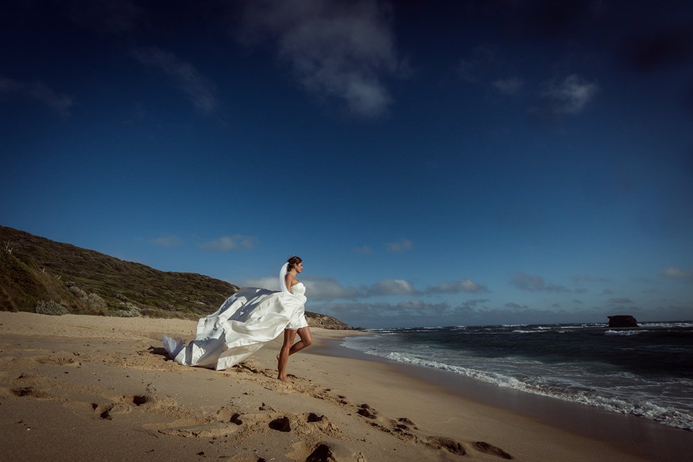 A woman in a wedding gown standing on a beach with the ocean and sky in the background.